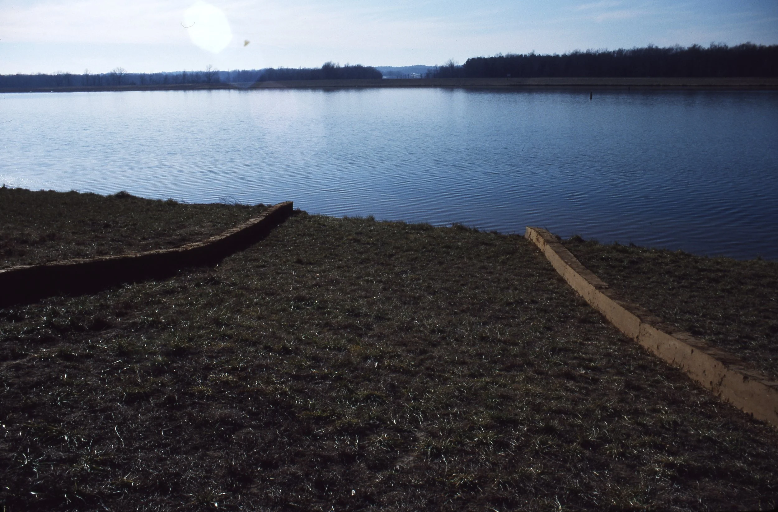 A lakeshore with a grassy bank, a curved wooden barrier, and calm water under a partly cloudy sky.