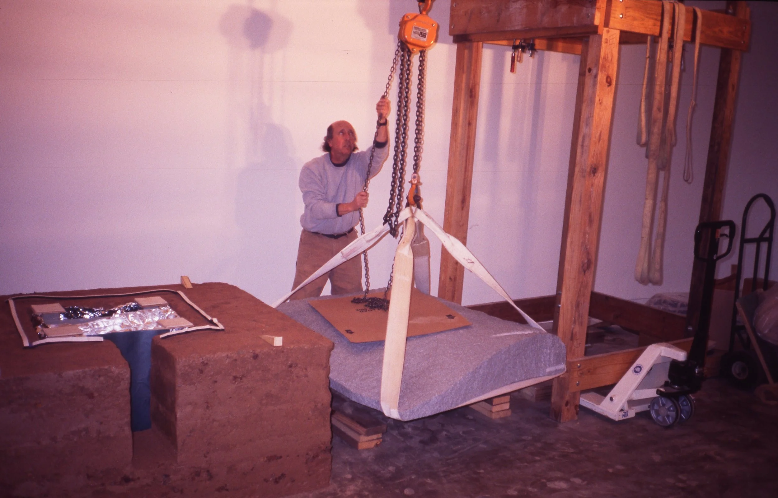 A man adjusts a large metal chain hoist attached to a wooden frame, with mummified or skeletal remains hanging from the chain. There are tools hanging on the wooden structure and a stone slab on a pallet, surrounded by straps and equipment.