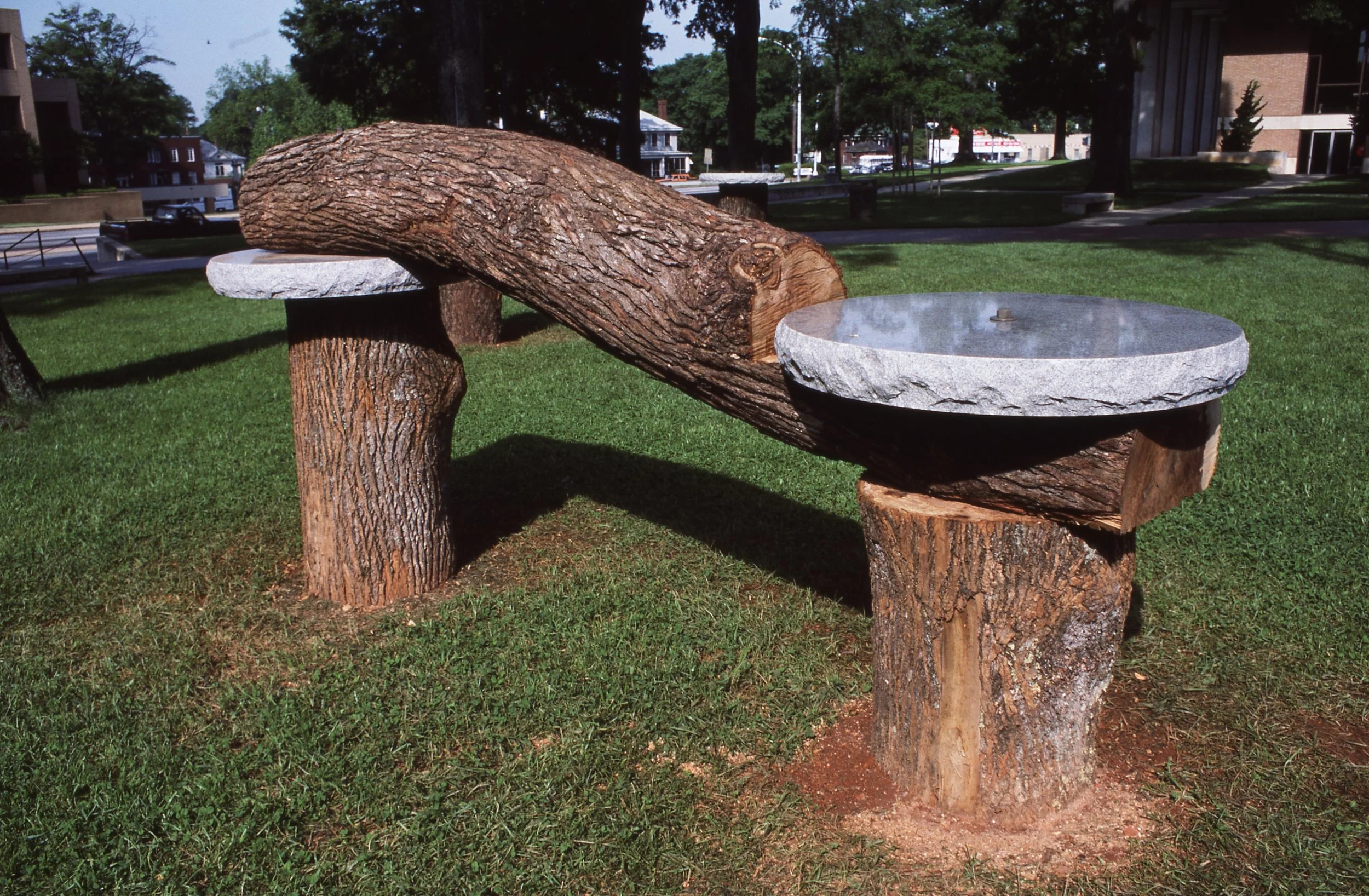 A park bench made from large logs with two circular stone tabletops, situated on a grassy area with trees and buildings in the background.