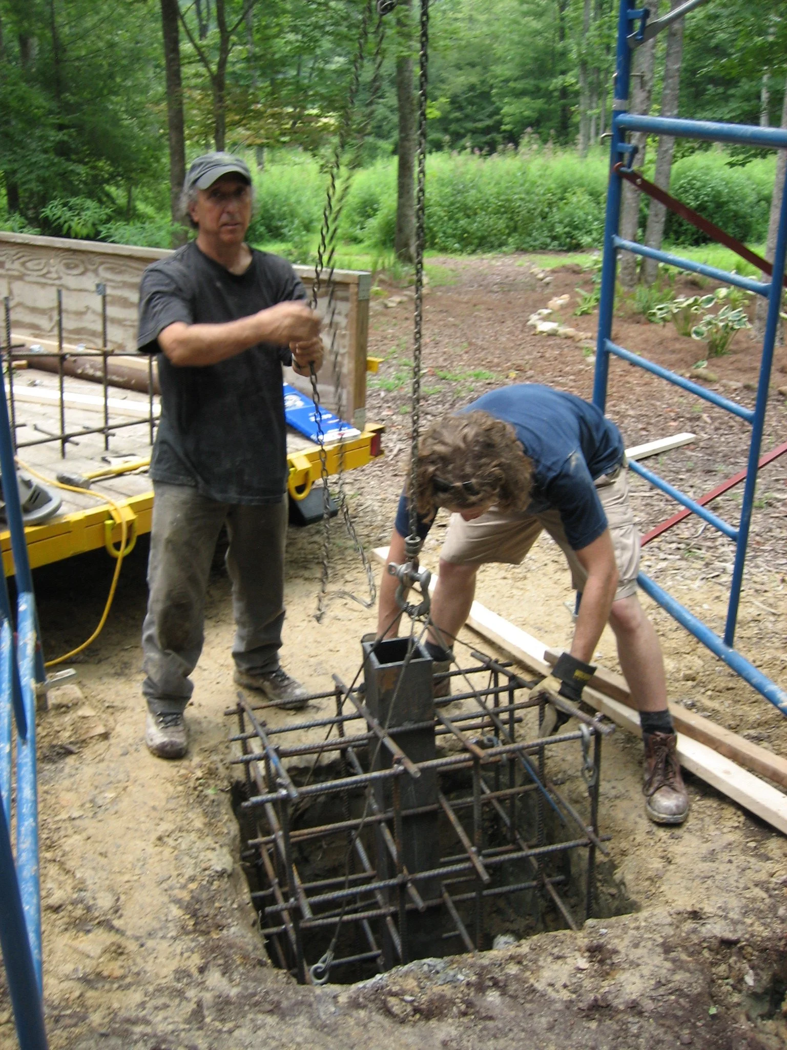Two people working on a construction project outdoors, with one man tightening a chain and another bending over a metal frame in a hole.