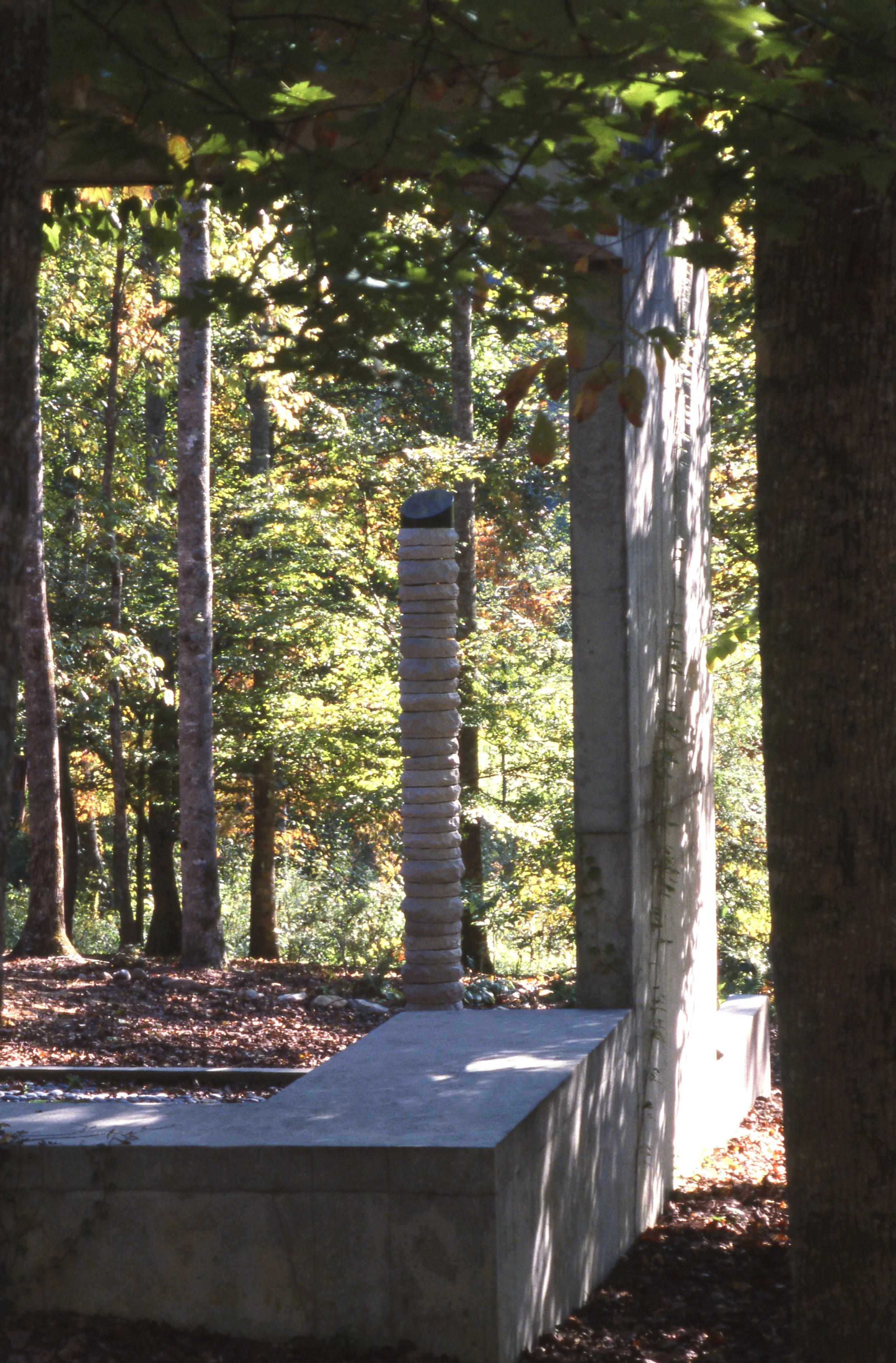 A stone sculpture situated outdoors amidst trees in a forest, with one tall, thin column of stacked stones and a curved concrete platform.