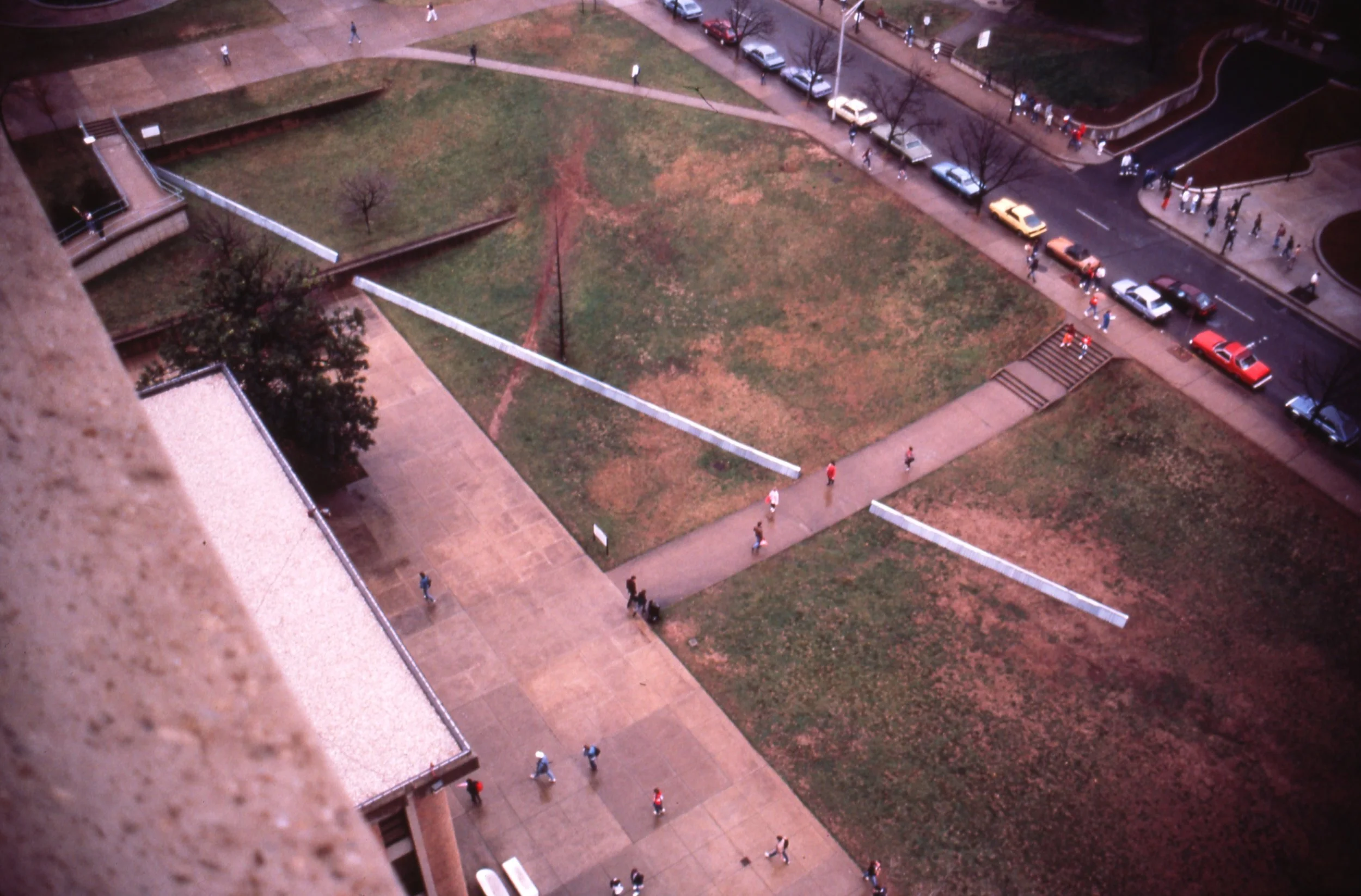 An aerial view of a city park with walkways, grassy areas, trees, and people walking. Parking lot with cars is visible along the street at the top of the image.