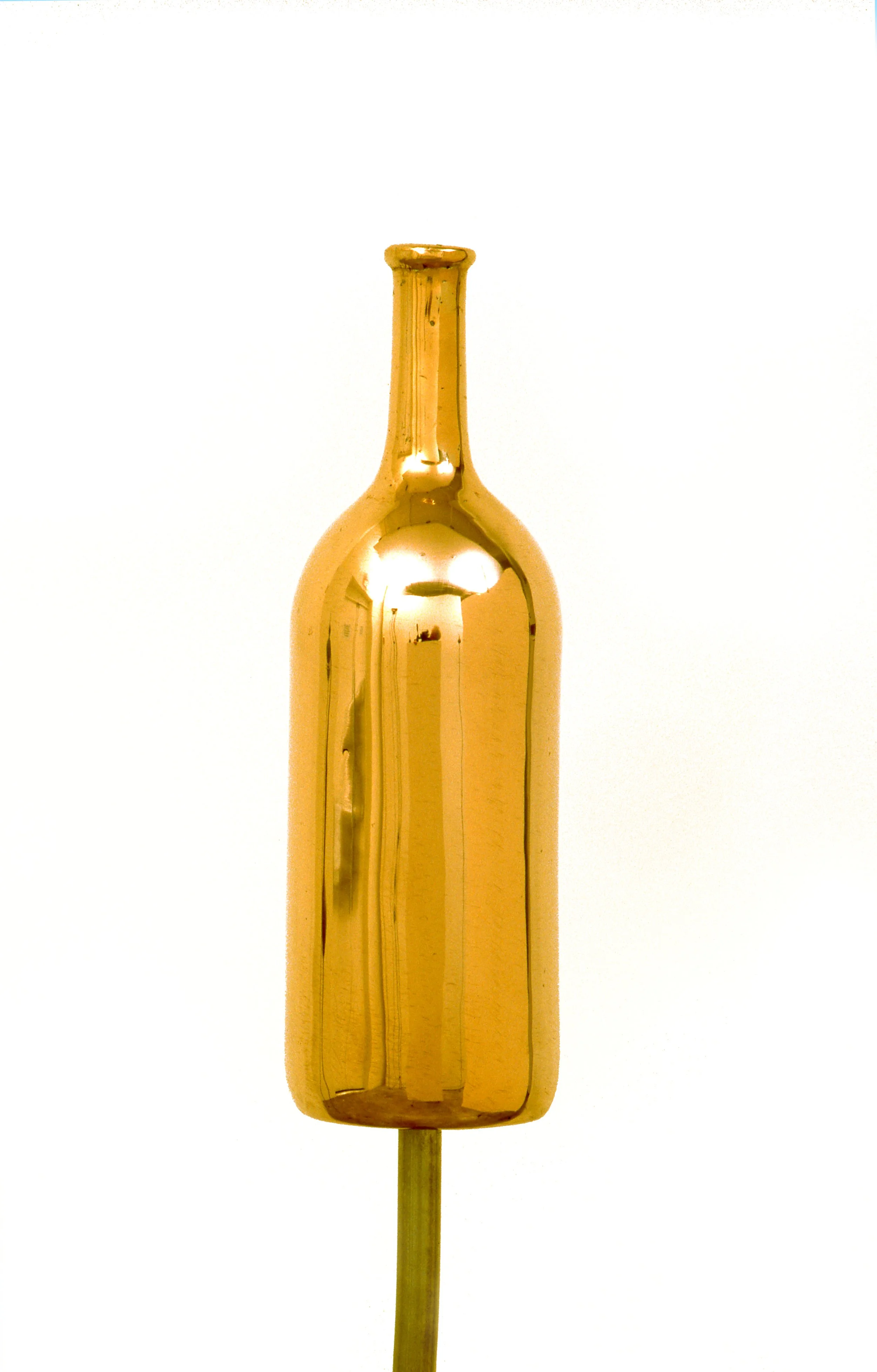 A gold-colored metallic bottle-shaped object mounted on a stick against a white background, resembling a decorative or artistic piece.