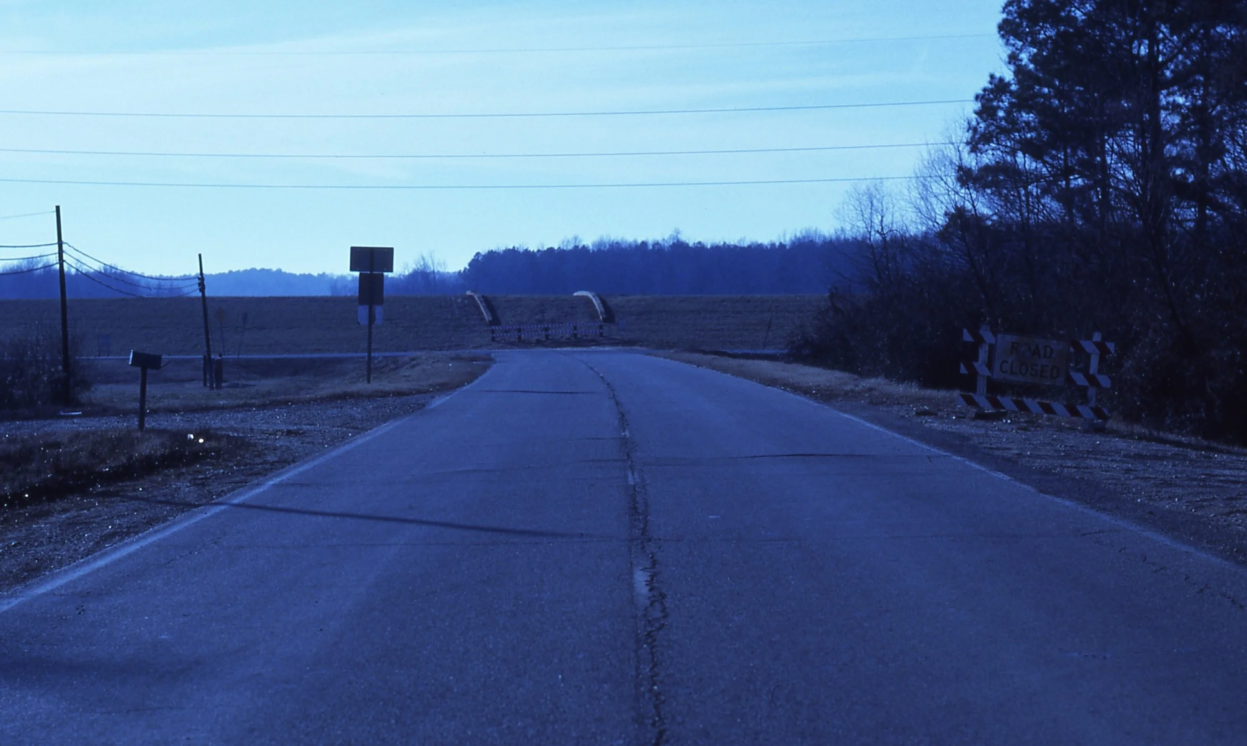 A deserted rural road with a road closed sign and barriers blocking the path, surrounded by trees and open fields under a blue sky.