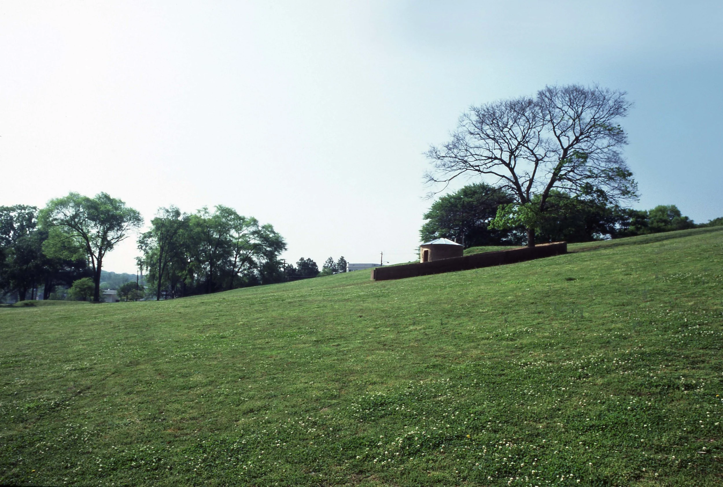 A park with lush green grass, several trees, and a small brick structure, with a bright sky in the background.