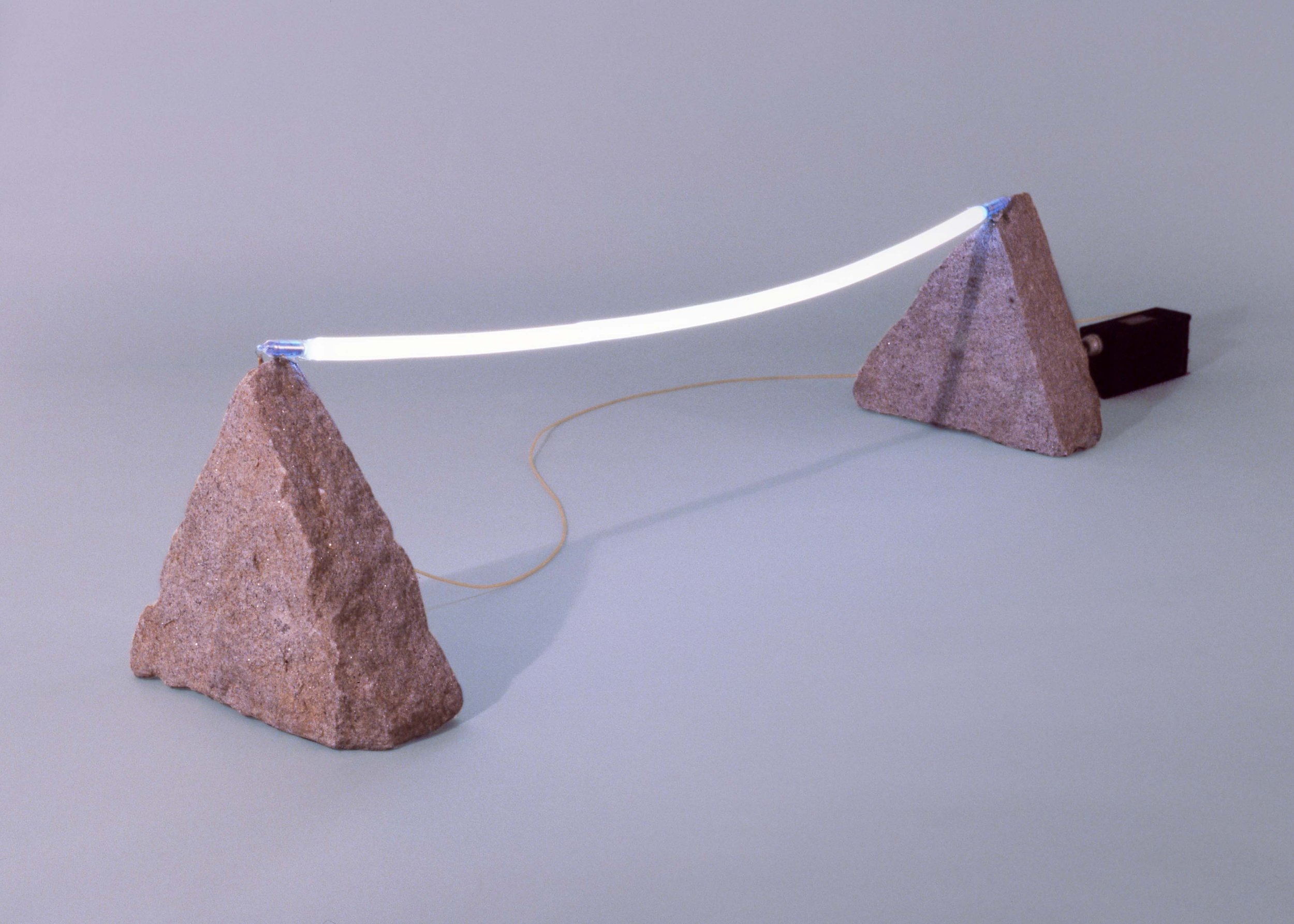 A scientific demonstration device with two large rocks supporting a glowing curved light beam, connected by a thin wire and powered by a small electric motor.