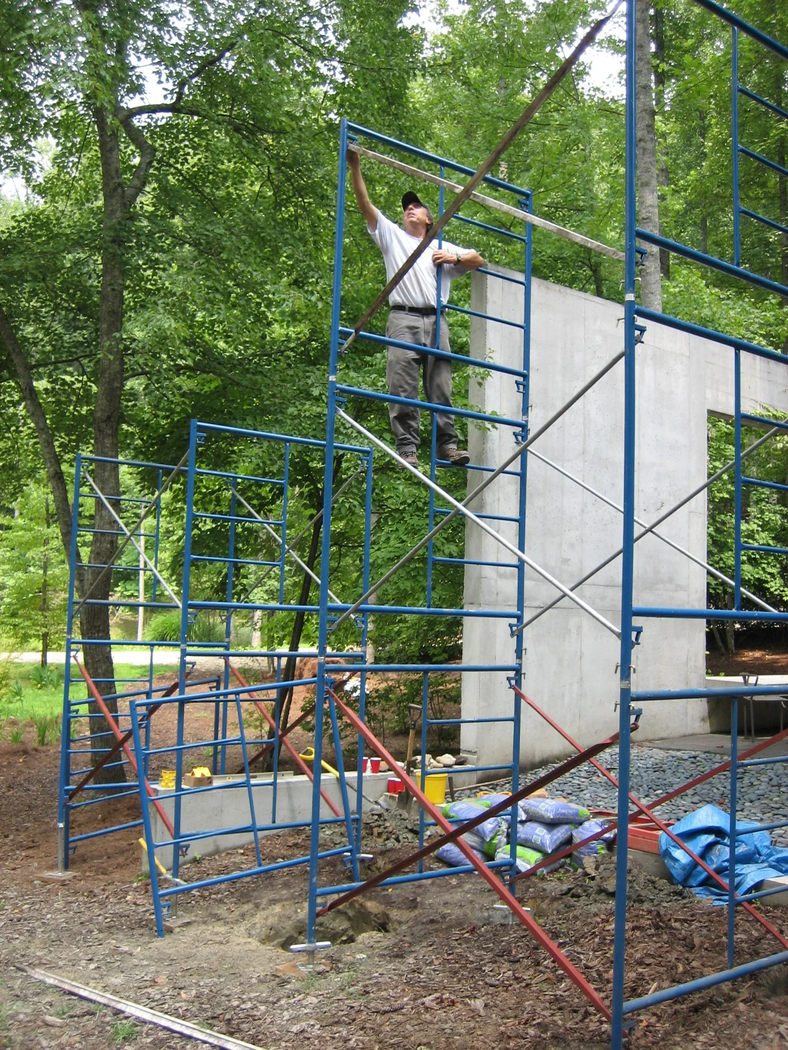 A man on a construction scaffold working on a concrete wall outdoors surrounded by green trees.
