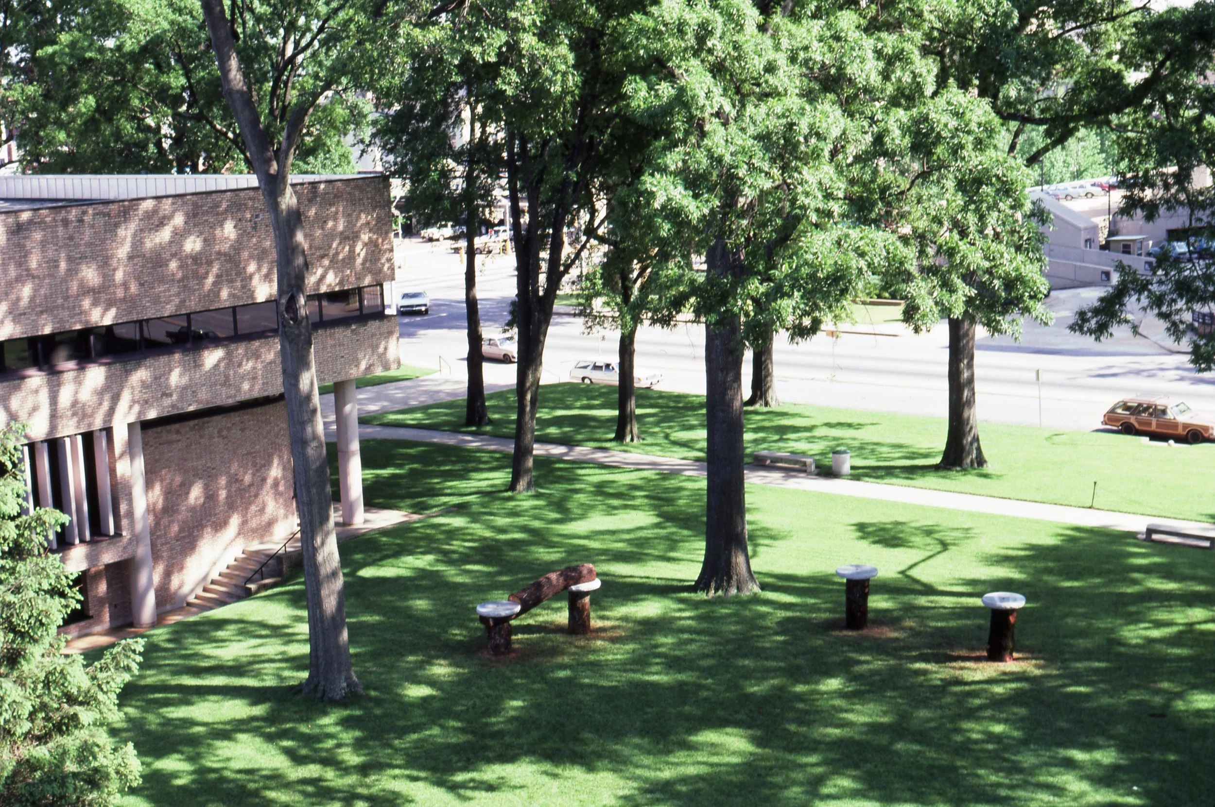 A park with green grass, tall trees, and benches surrounded by a brick building and a road with parked cars in the background.