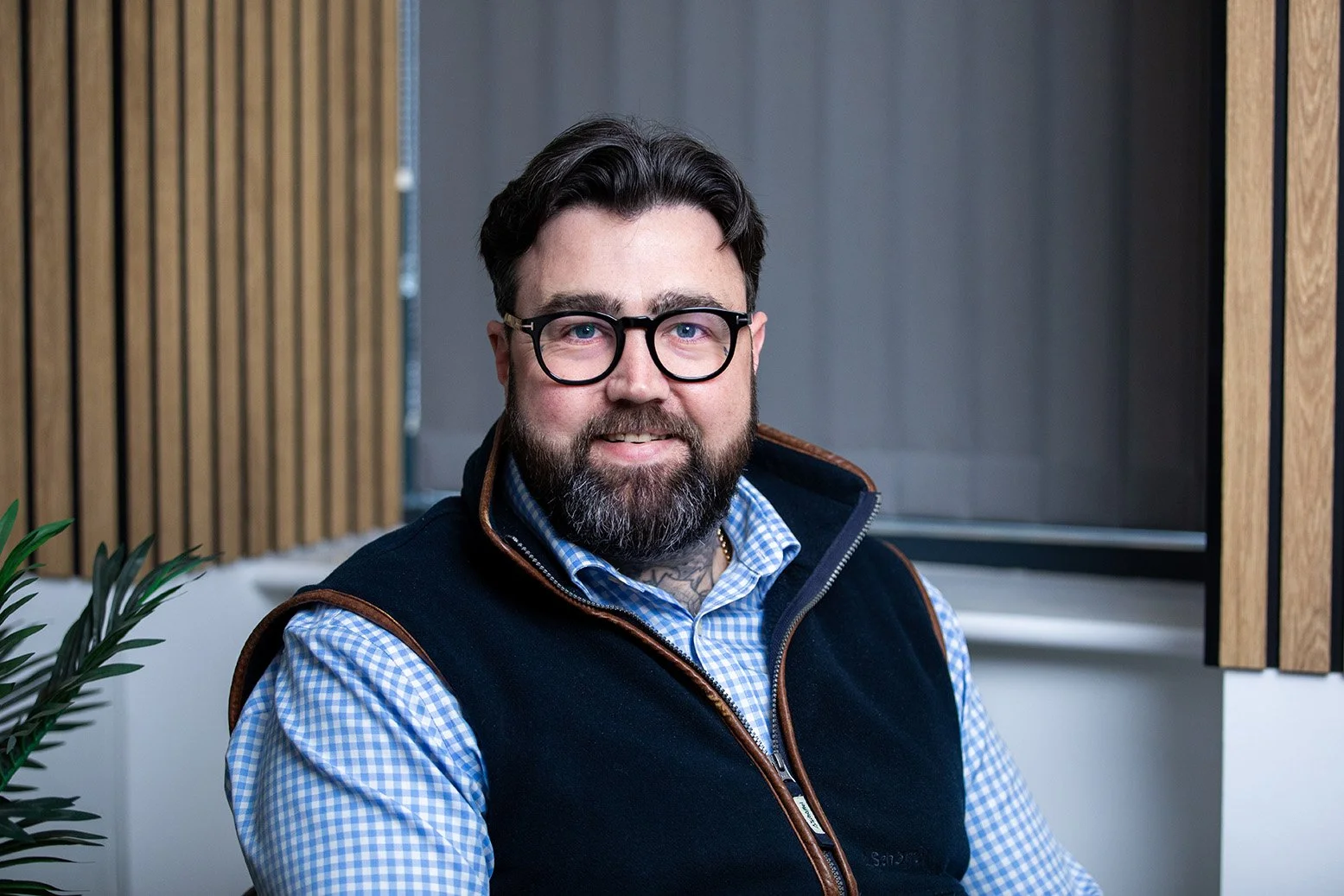 Headshot image of a white man with glasses. Wearing a check blue and white shirt and a navy gilet.