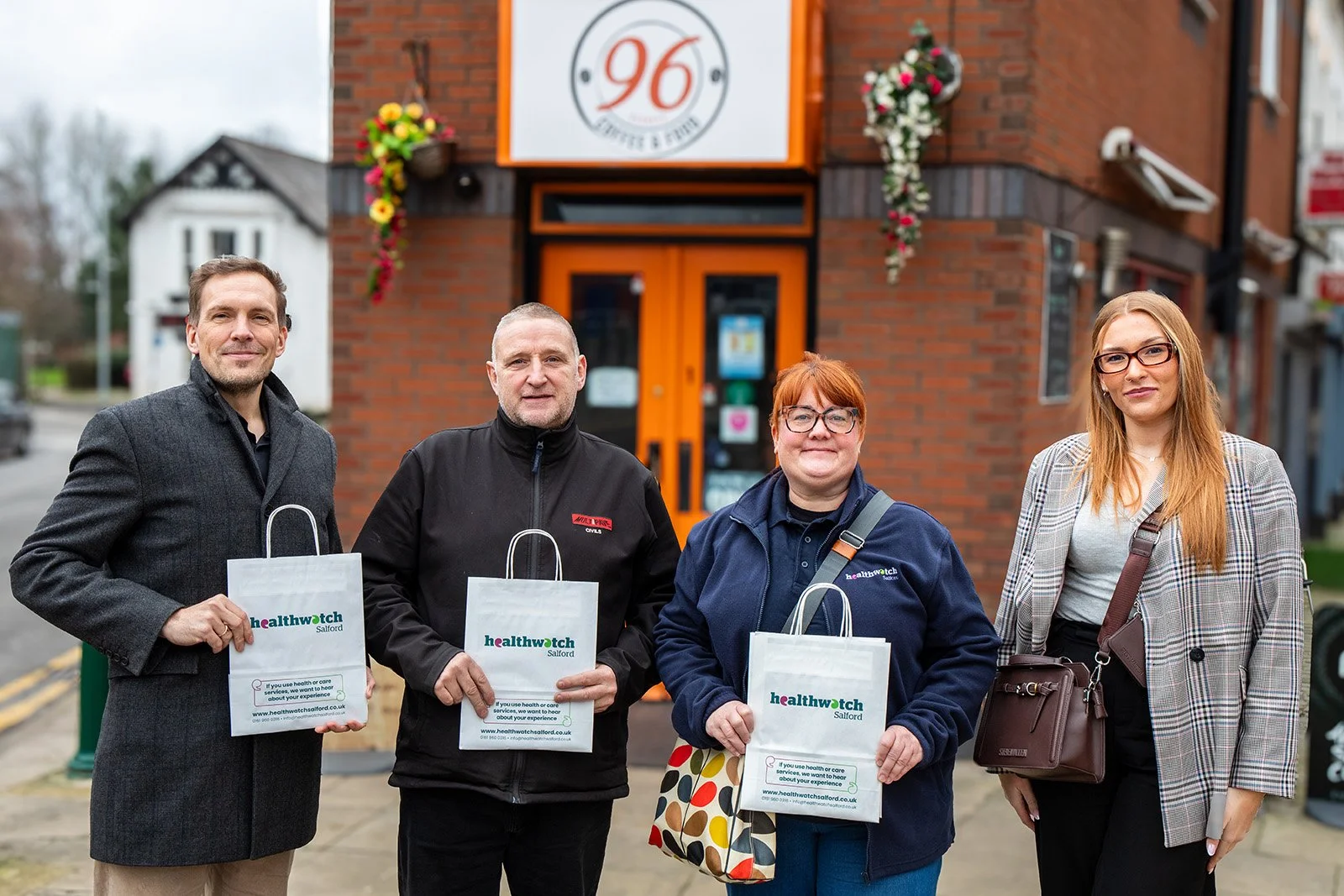 Two white males and two white females standing outside a cafe holding Healthwatch Salford bags