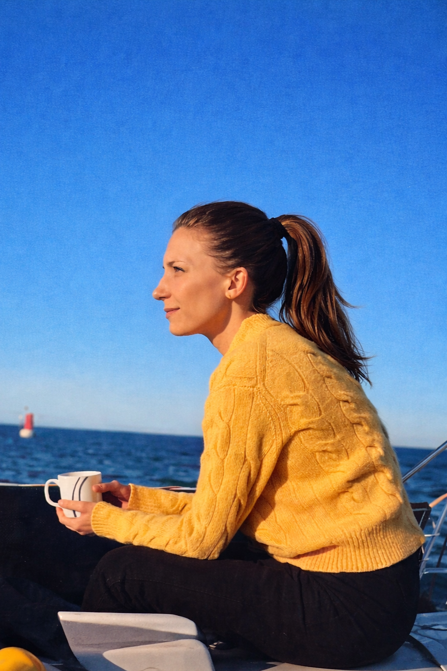 Femme assise sur un bateau, tenant une tasse, avec la mer et un phare en arrière-plan, sous un ciel clair.