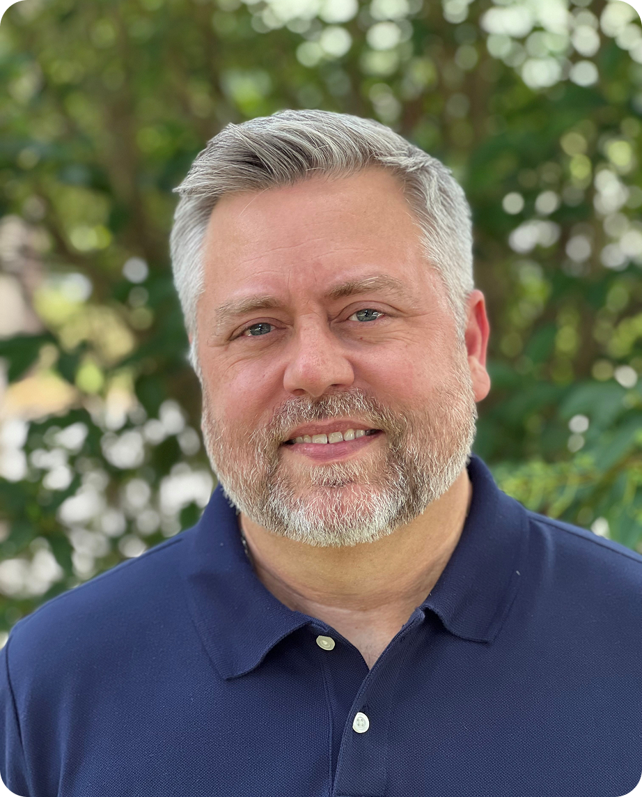 A middle-aged man with gray hair and beard smiling outdoors, wearing a navy blue polo shirt, with green trees in the background.