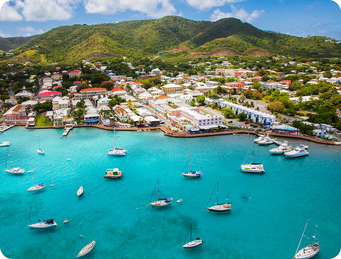 Aerial view of a coastal town with colorful buildings, boats in turquoise water, and green hills in the background.