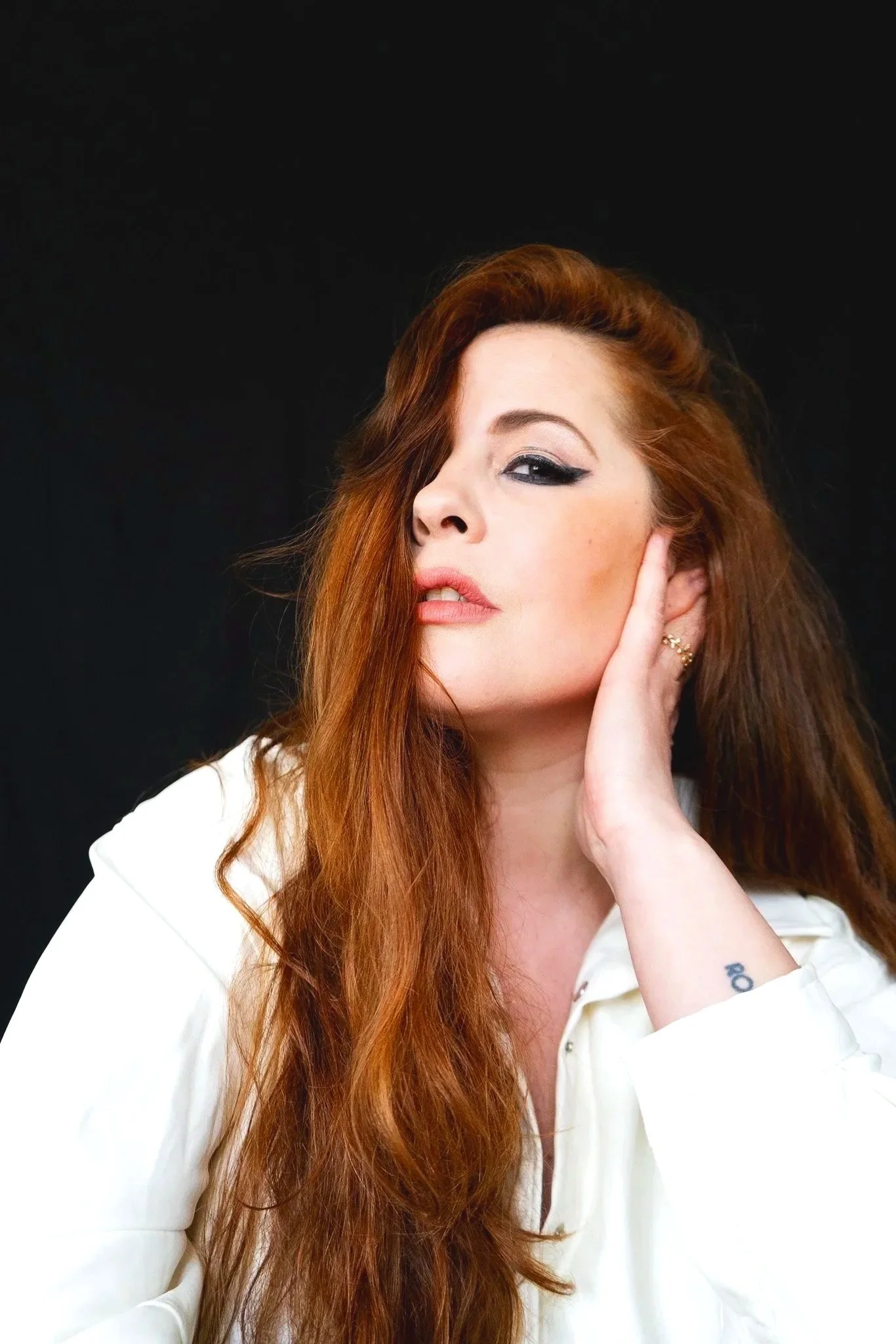 Femme aux cheveux longs et roux, portant une chemise blanche, posant avec une main près de l'oreille sur un fond noir.