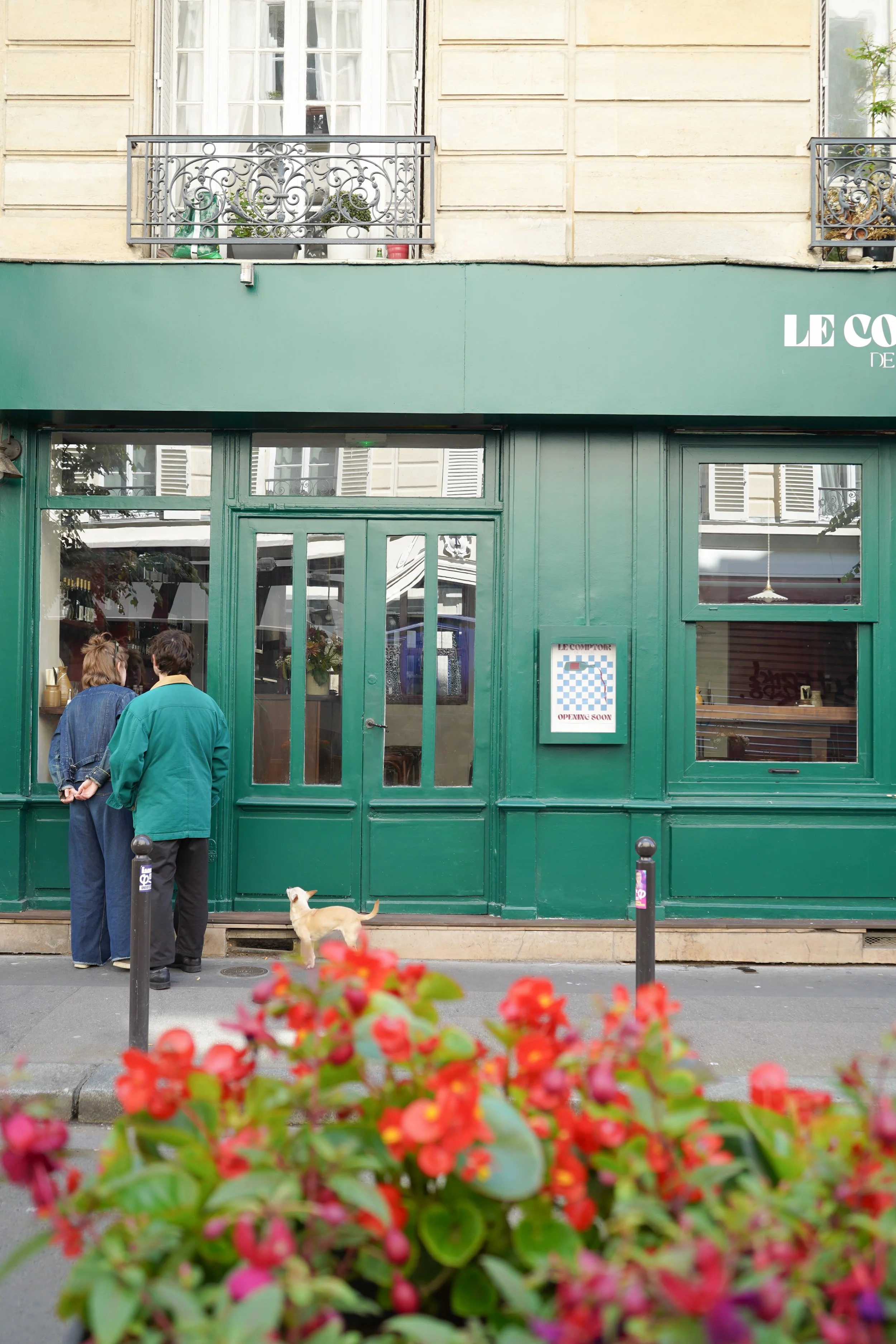 Deux personnes discutent devant un restaurant vert avec une enseigne indiquant qu'il ouvre bientôt. Un chien se tient à côté d'eux, et des fleurs rouges floues sont au premier plan.