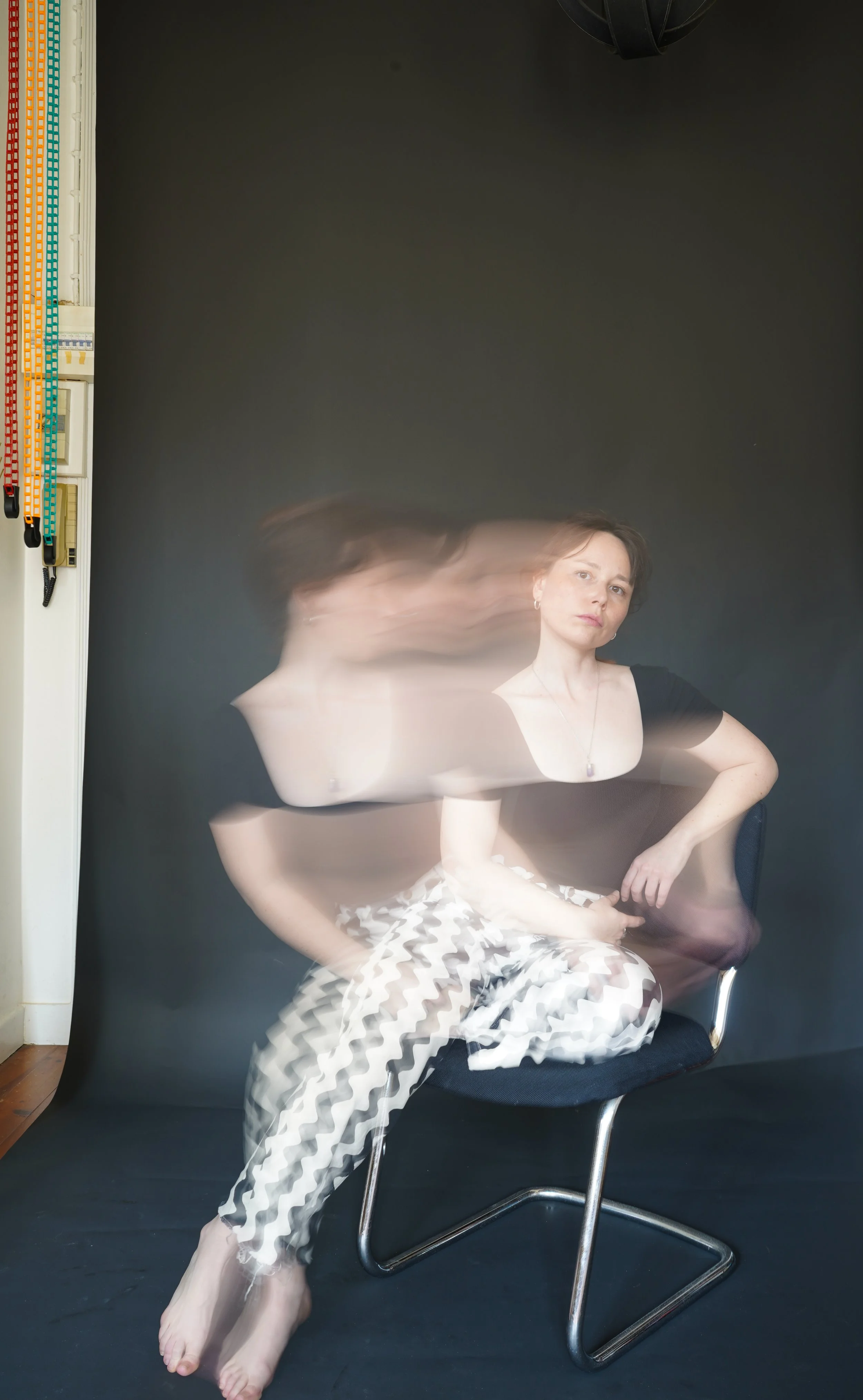 Une femme assise sur une chaise noire, mouvement flou indiquant un effet de longue exposition, avec un fond noir