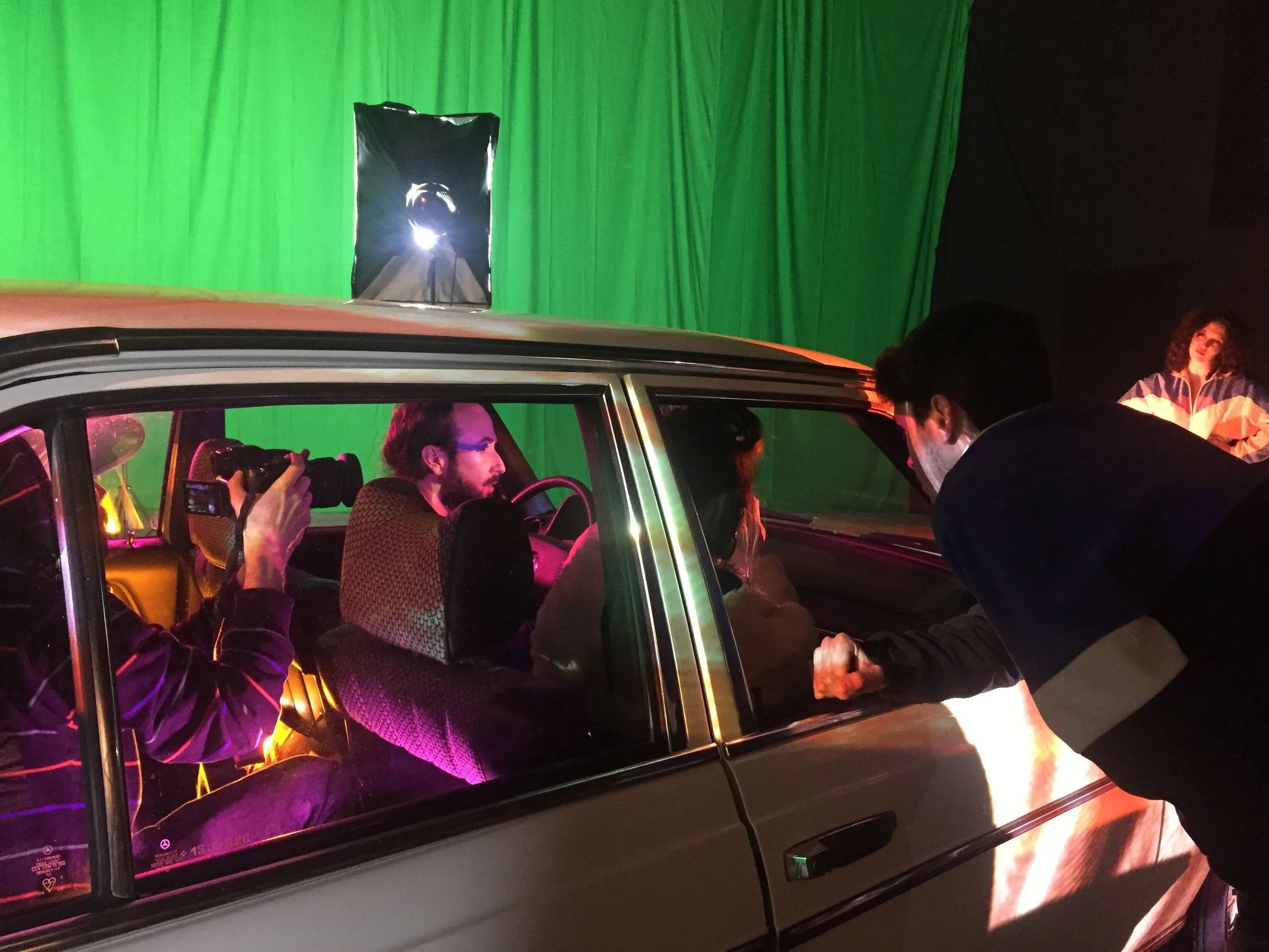 Une séance de tournage filmée dans un studio avec un fond vert, plusieurs personnes autour d'une voiture, dont une personne dans la voiture parlant avec quelqu'un à l'extérieur, une personne avec un appareil photo, et un éclairage professionnel.