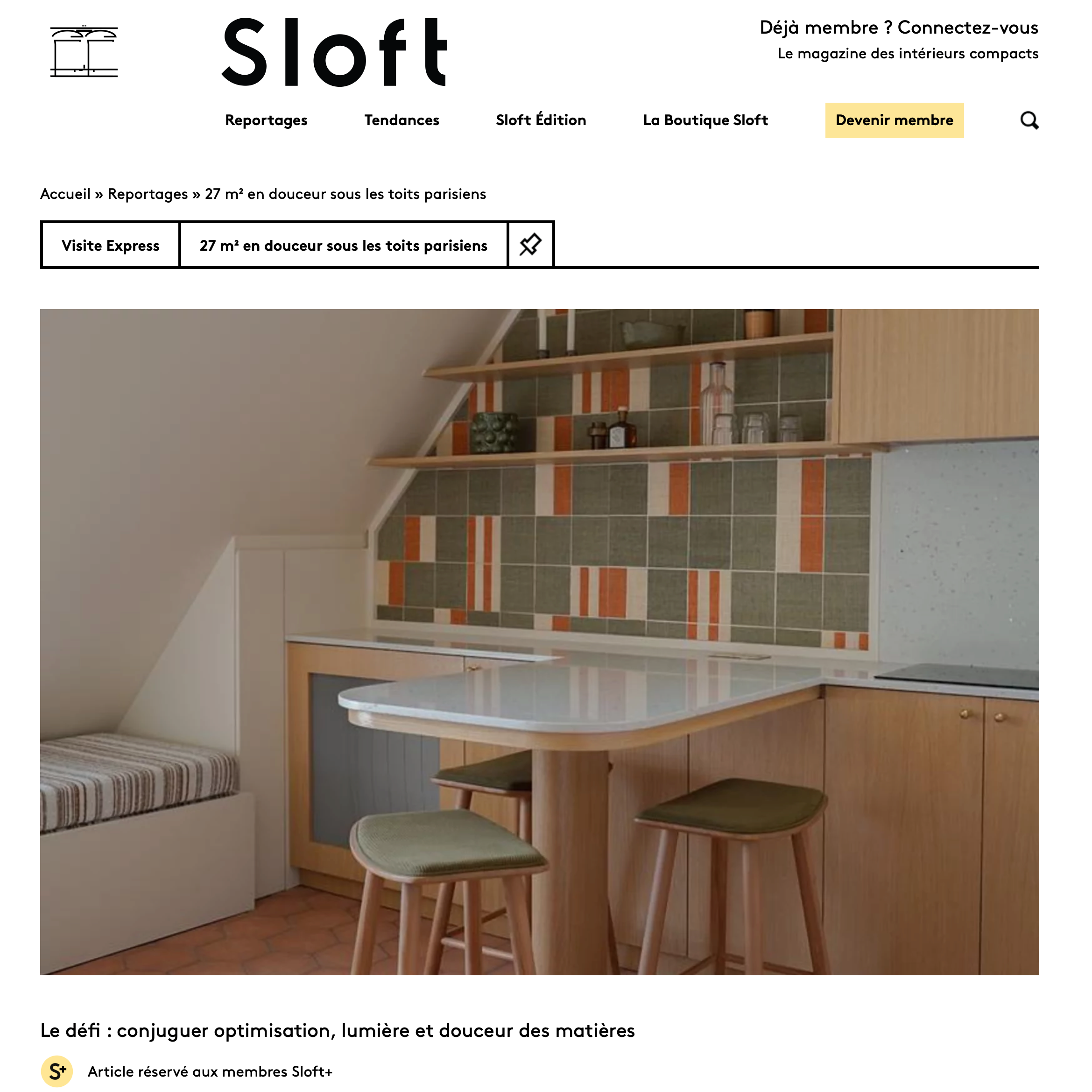 Intérieur d'une cuisine avec une table en formica blanc, des tabourets en bois avec des coussins verts, meubles en bois clair, étagères ouvertes en bois avec quelques objets, murs décorés avec des carreaux colorés vert, orange et blanc.