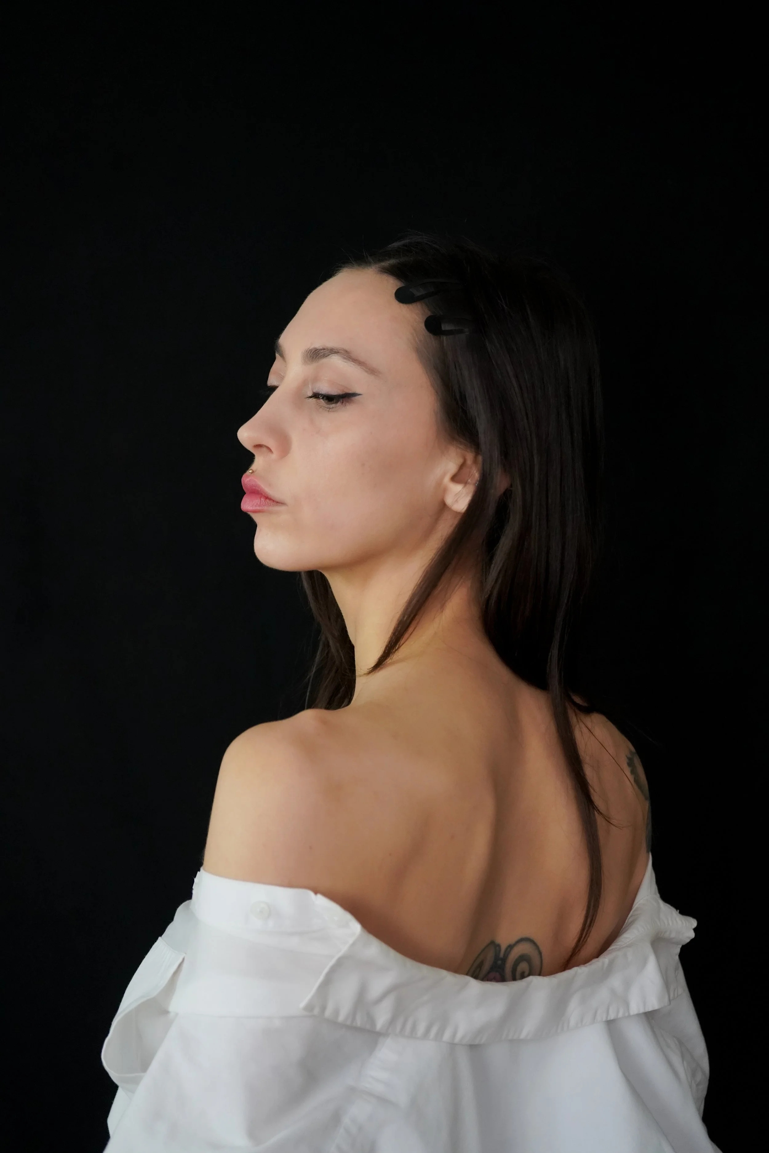 Jeune femme avec maquillage discret et tatouages, portant une robe blanche décontractée, vue de profil contre un fond noir.