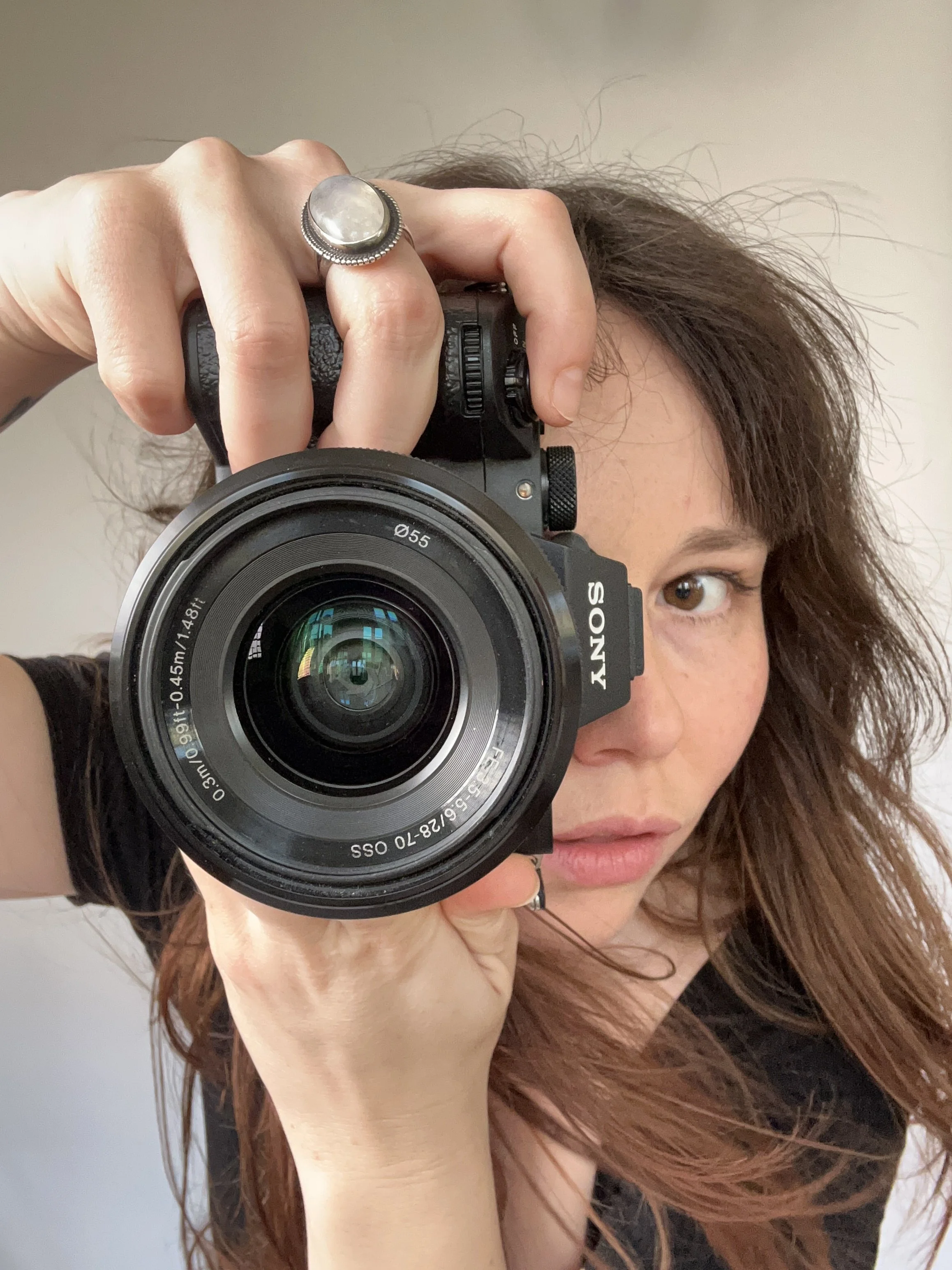 Une femme avec des cheveux ondulés bruns, portant un anneau sur le doigt, prend une photo avec un appareil photo Sony. L'objectif de l'appareil est pointé vers l'objectif.