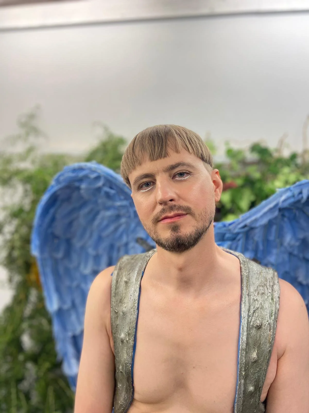 Homme avec des ailes bleues, portant un sac à dos argenté, posant devant un fond de plantes vertes.