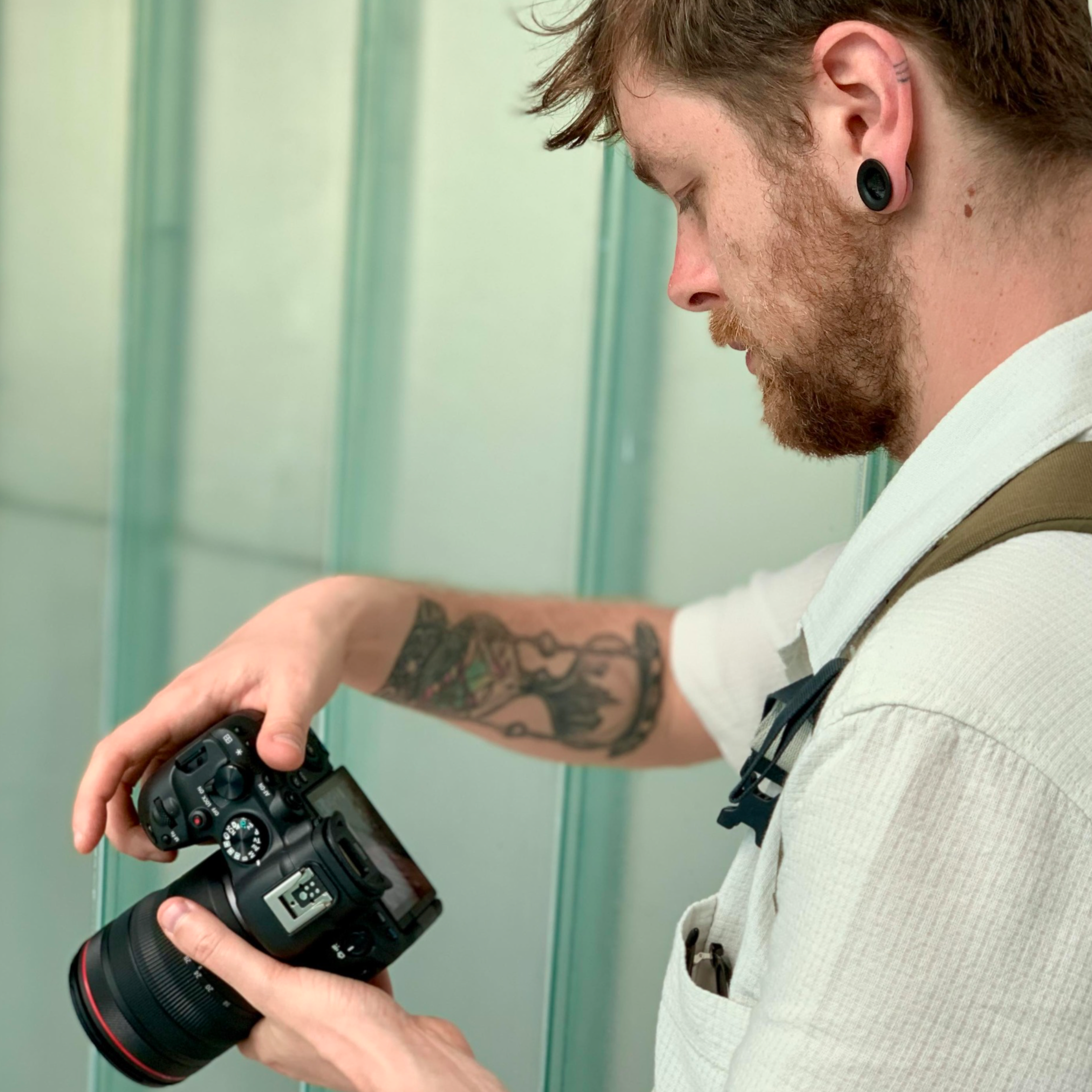 Un homme avec une barbe et un tatouage au bras, portant une chemise blanche, regarde un appareil photo numérique en main dans un environnement avec des murs verts.