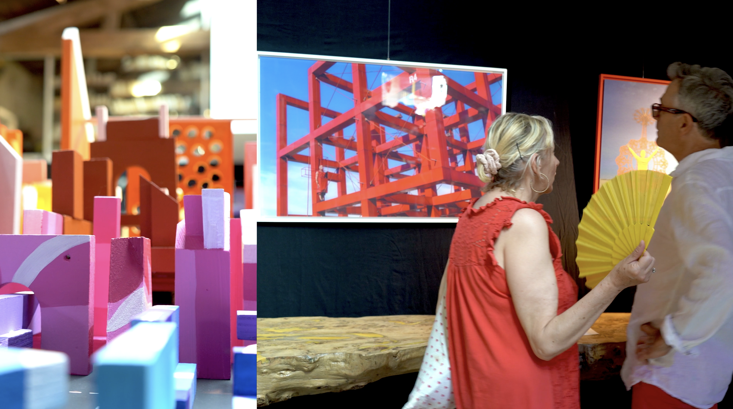 Deux personnes discutent dans une galerie d'art, un homme avec des lunettes et une femme portant un fan jaune, entourés d'œuvres d'art modernes. Sur un écran derrière eux, une structure rouge en bois