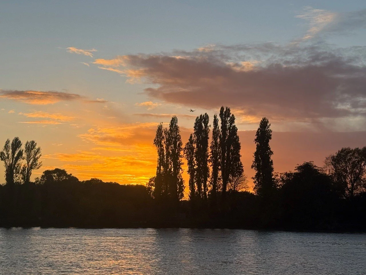 Sunset over a river with tall trees along the shoreline and an airplane flying in the sky.