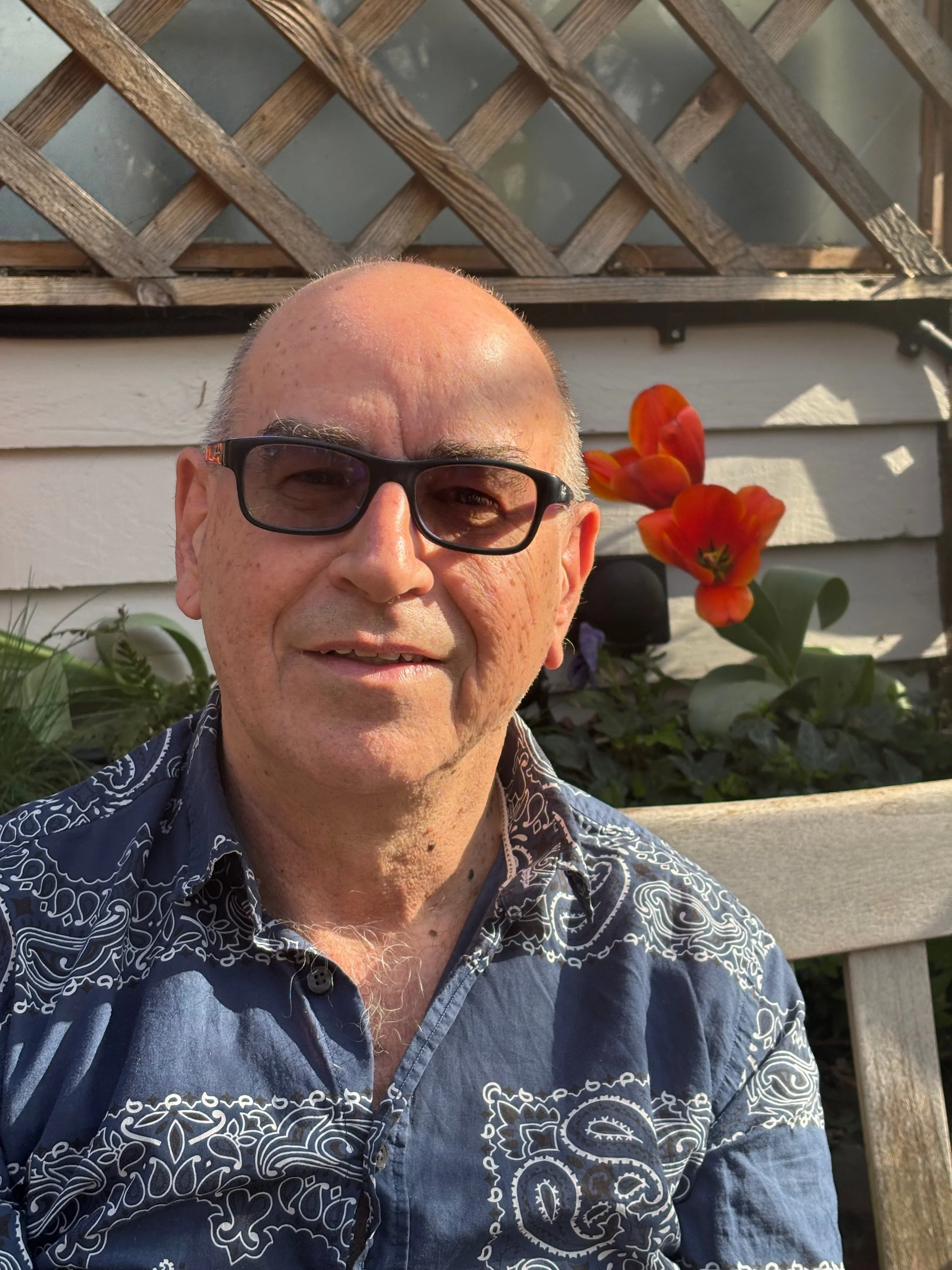 A man wearing glasses and a patterned blue shirt is smiling outdoors in a garden with orange flowers and greenery behind him.