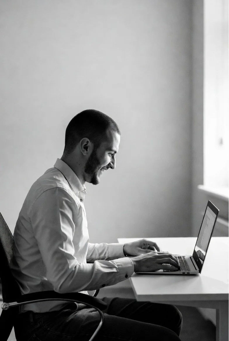 A man sitting at a desk using a laptop, smiling and looking at the screen, in a well-lit room.