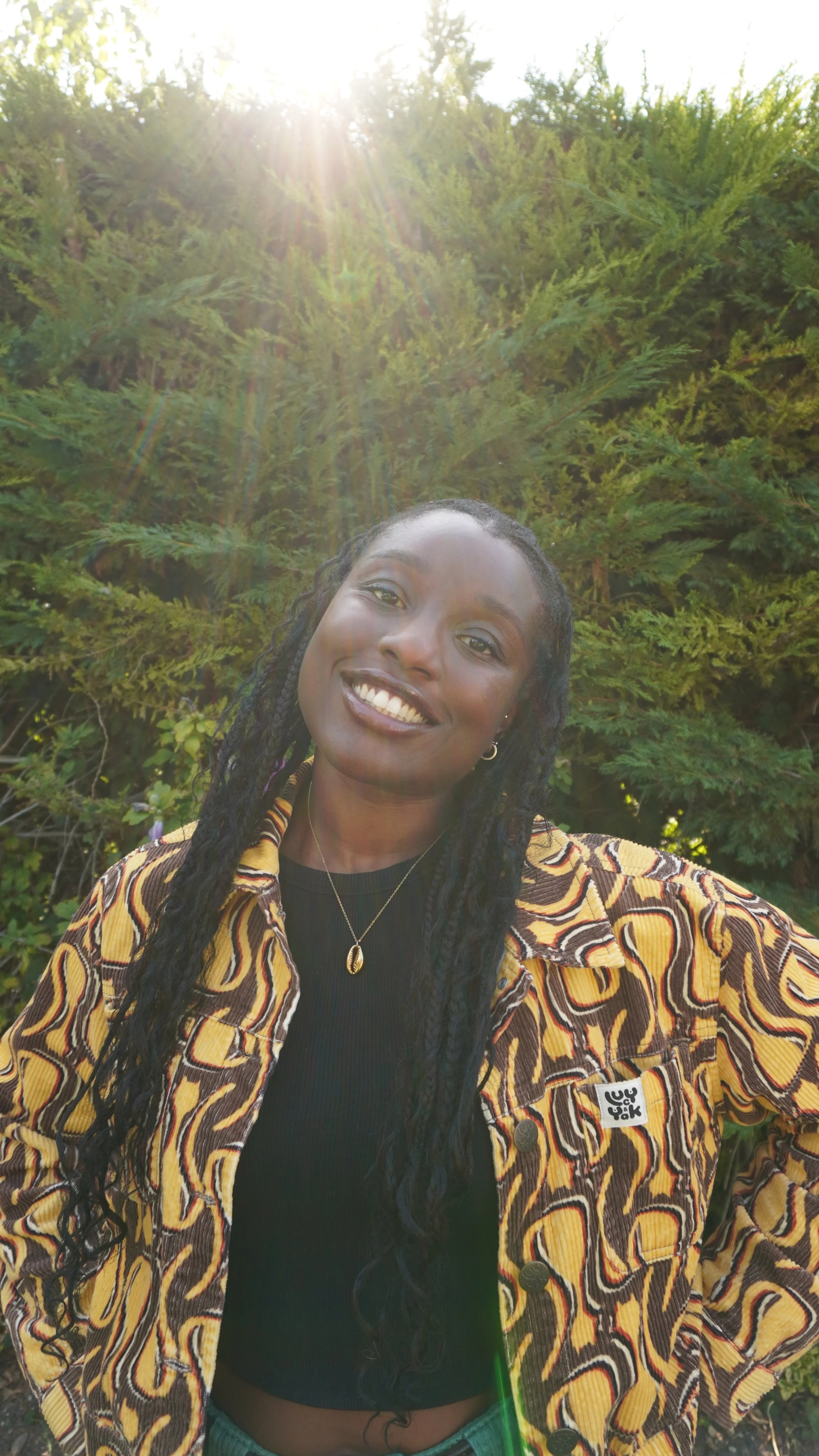 A woman taking a selfie outdoors with green trees in the background, sunlight shining down, wearing a patterned jacket and smiling.