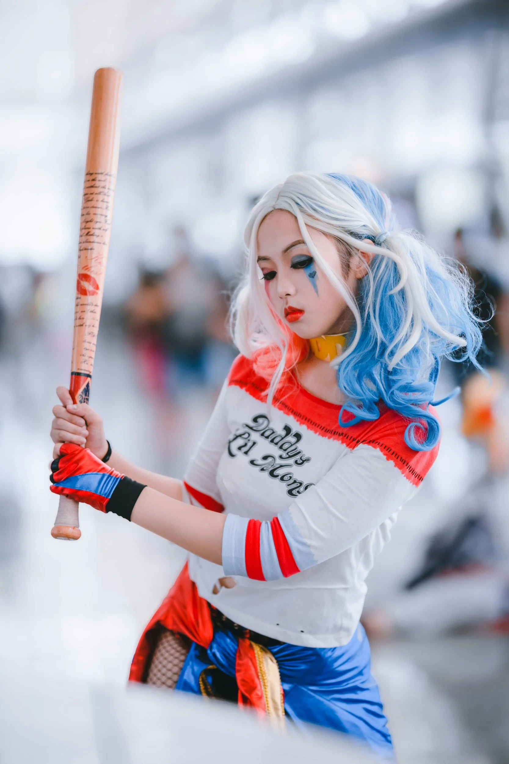 A woman dressed as Harley Quinn with blue and pink hair, wearing a white t-shirt that says "Daddy's Lil Monster," holding a baseball bat, at a cosplay event.