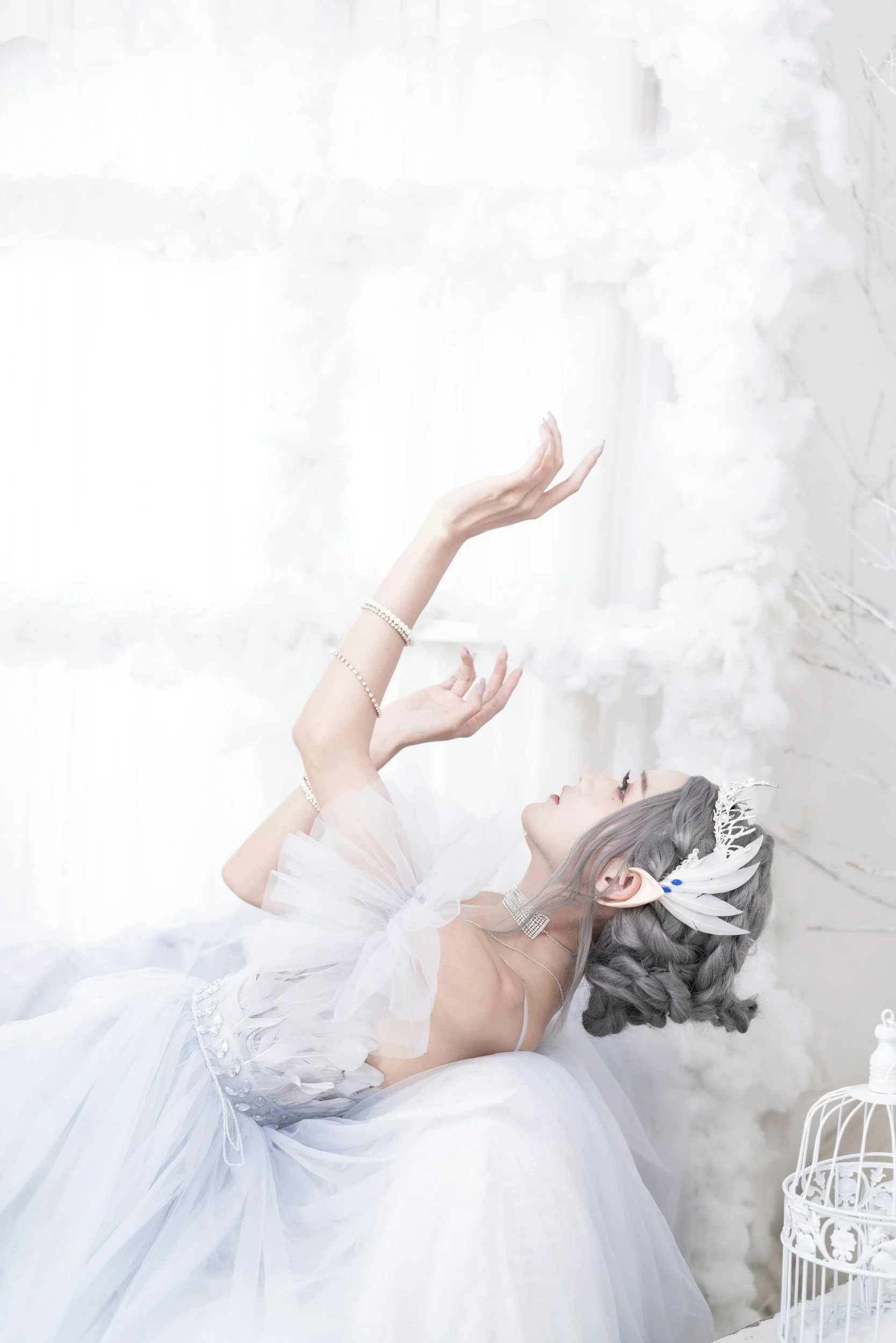 A woman wearing a white dress with tulle and pearl jewelry, laying on a white surface with her eyes closed and hands raised, in a bright, airy room decorated with white flowers and a birdcage.