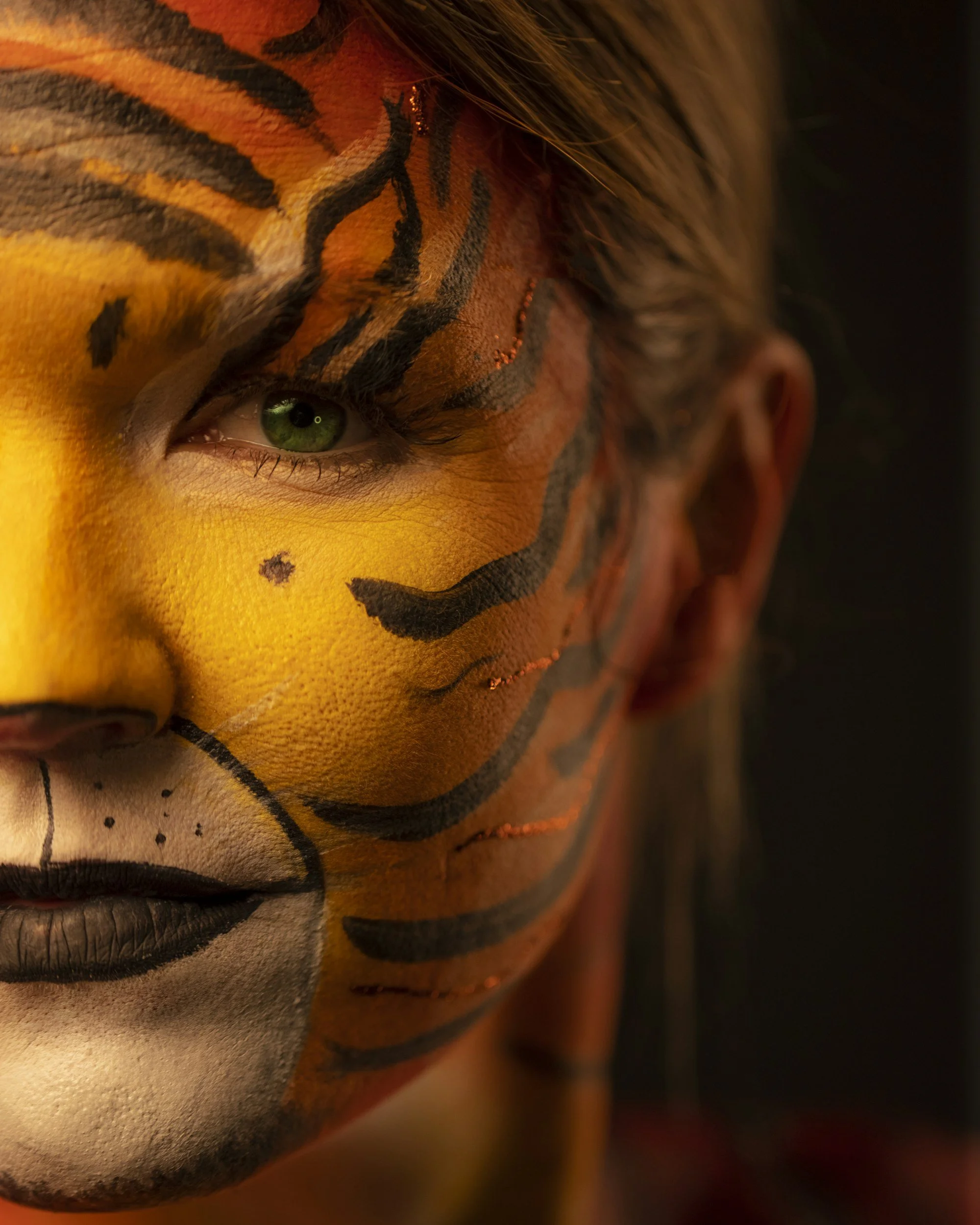 Close-up of a person's face with tiger face paint, featuring yellow, black, and white colors, and green eyes.