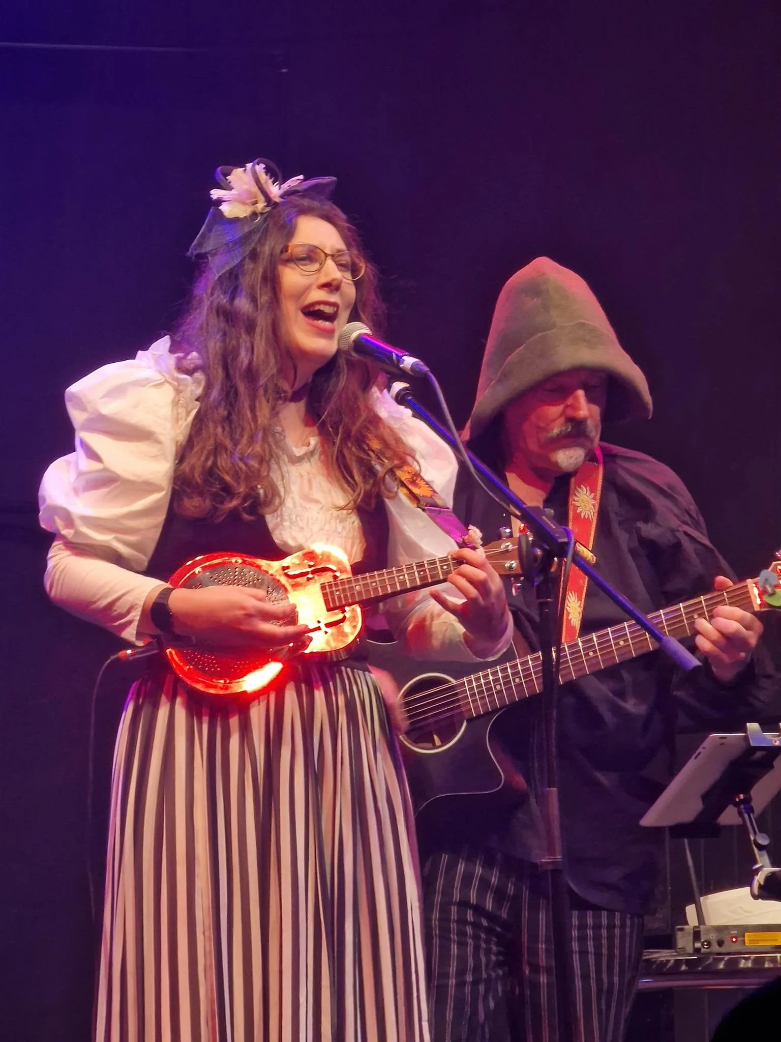 Two musicians performing on stage: a woman singing into a microphone while playing a ukulele, and a man playing an acoustic guitar wearing a large hat.