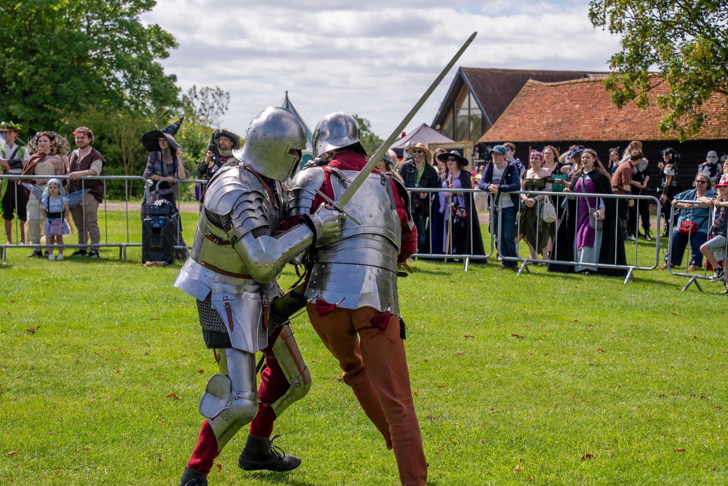 Two people dressed in medieval armor engaged in a mock sword fight in a grassy field during daylight.
