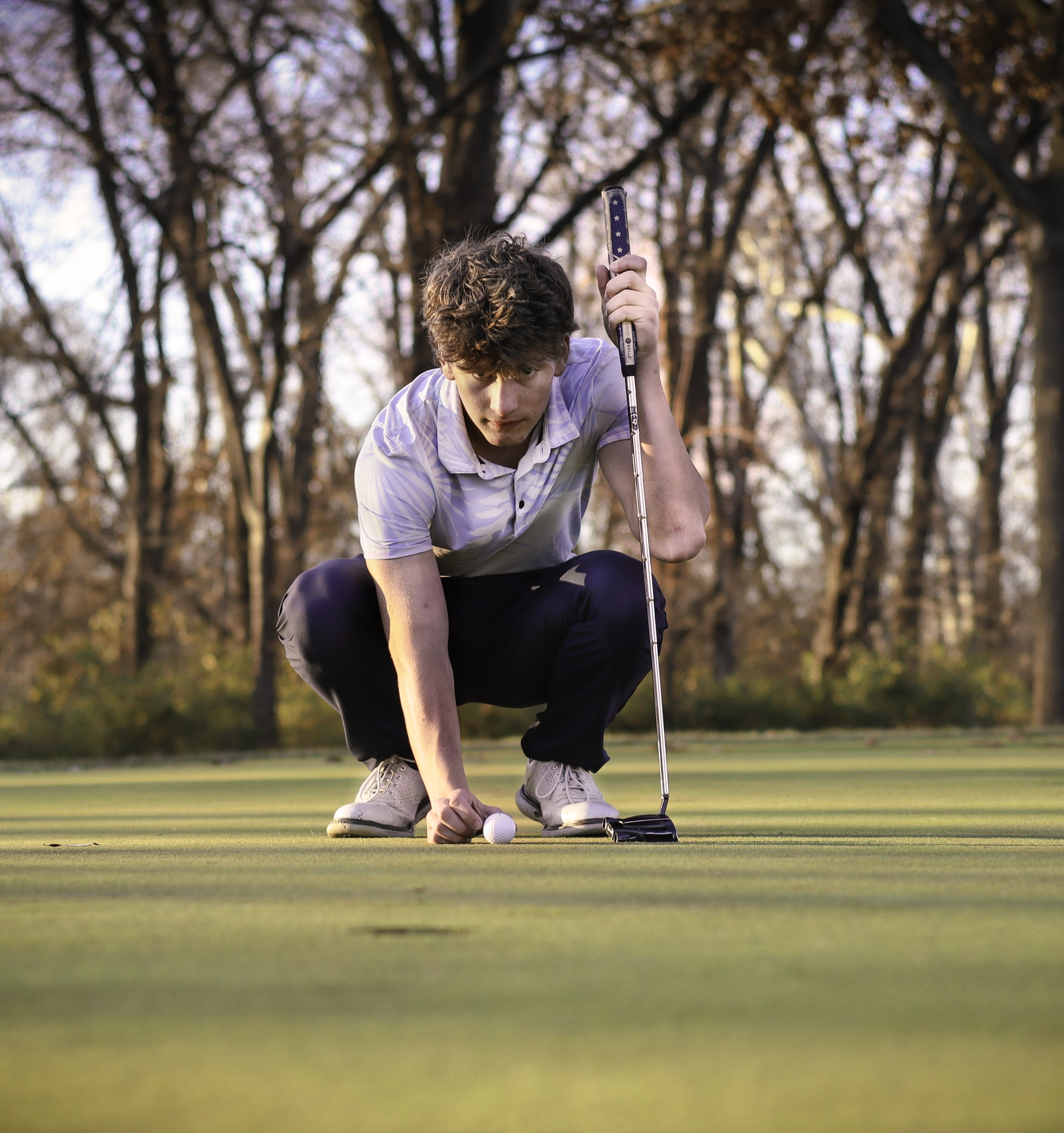 A young man on a golf course crouching down to line up a shot, holding a golf club, with a golf ball on the green in front of him, surrounded by trees with autumn leaves.