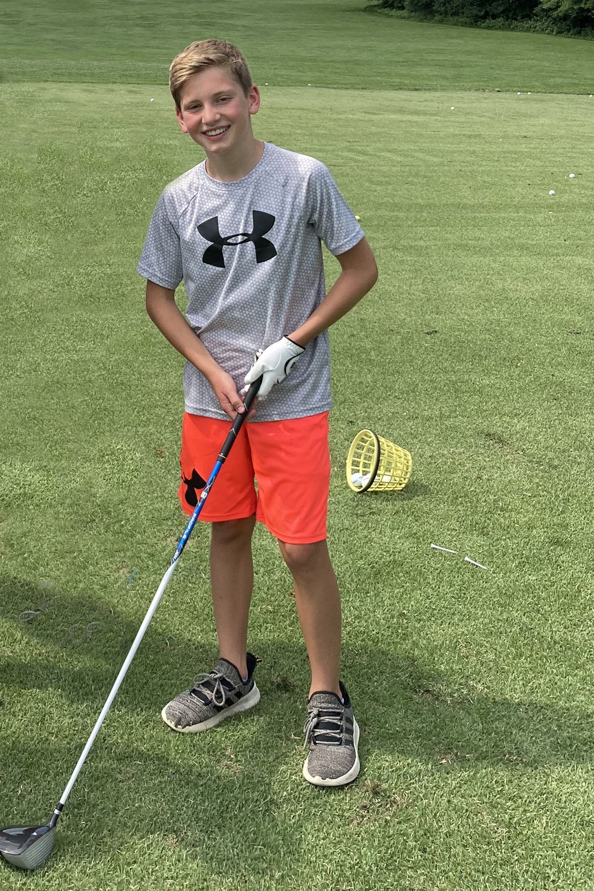 Young boy holding a golf club on a golf course with a yellow basket near his feet and a golf ball in it.