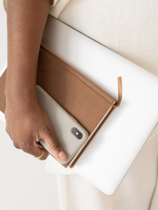 Person holding a smartphone with a brown notebook on top, walking with a white laptop in their hand.