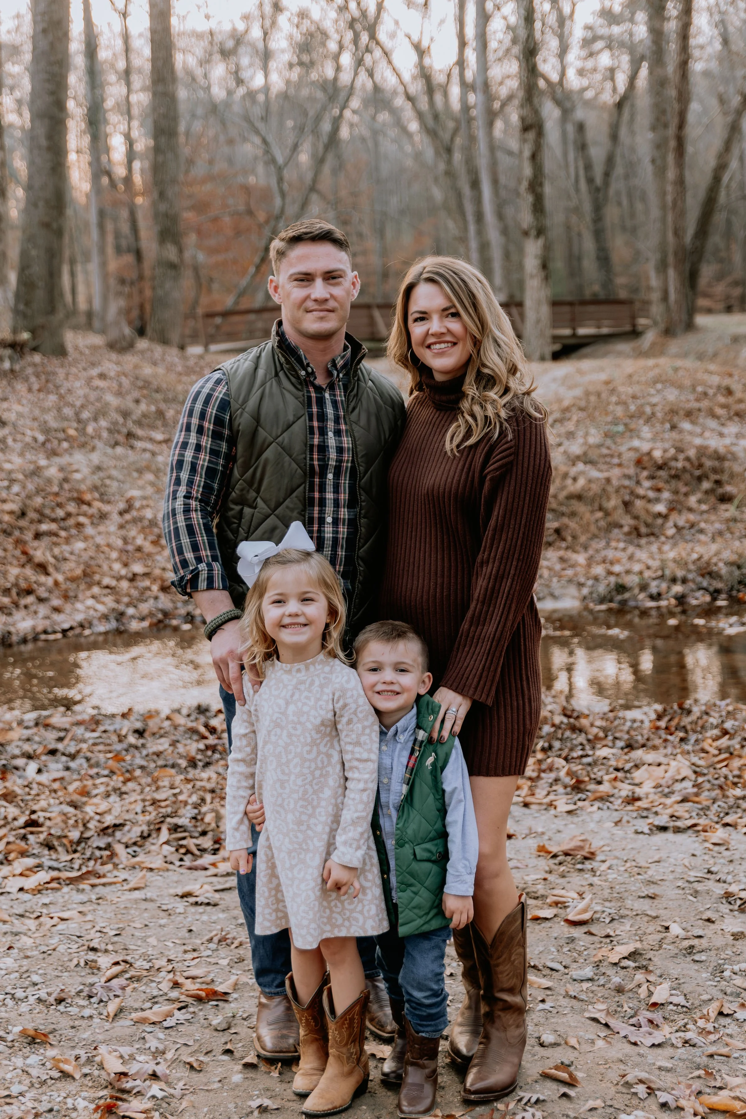 A family of four standing outdoors in a wooded area during autumn, with fallen leaves on the ground and a small creek behind them.