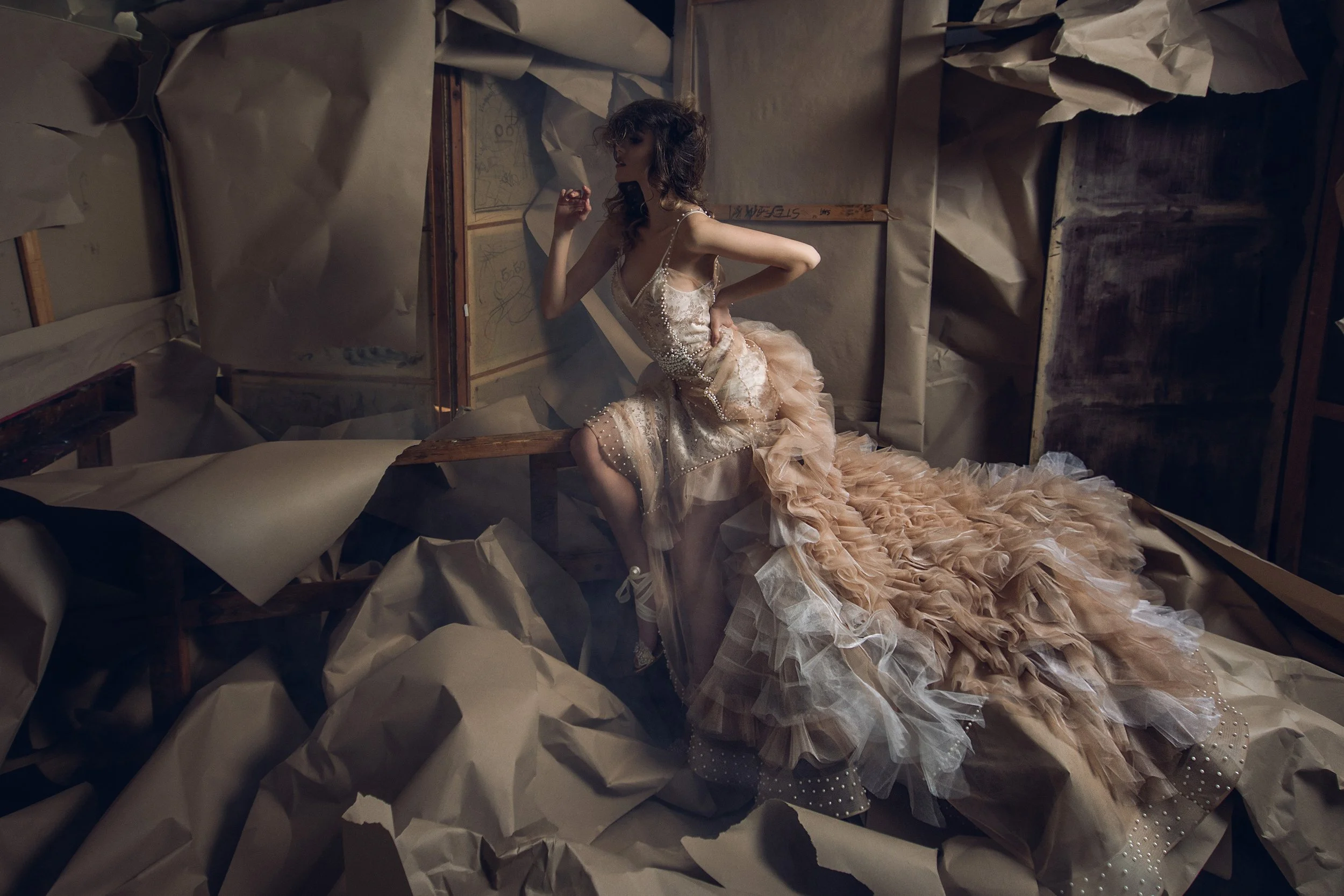 A woman in a beige, ruffled, floor-length gown with pearl embellishments sitting amidst crumpled paper on the floor and painted walls in the background.