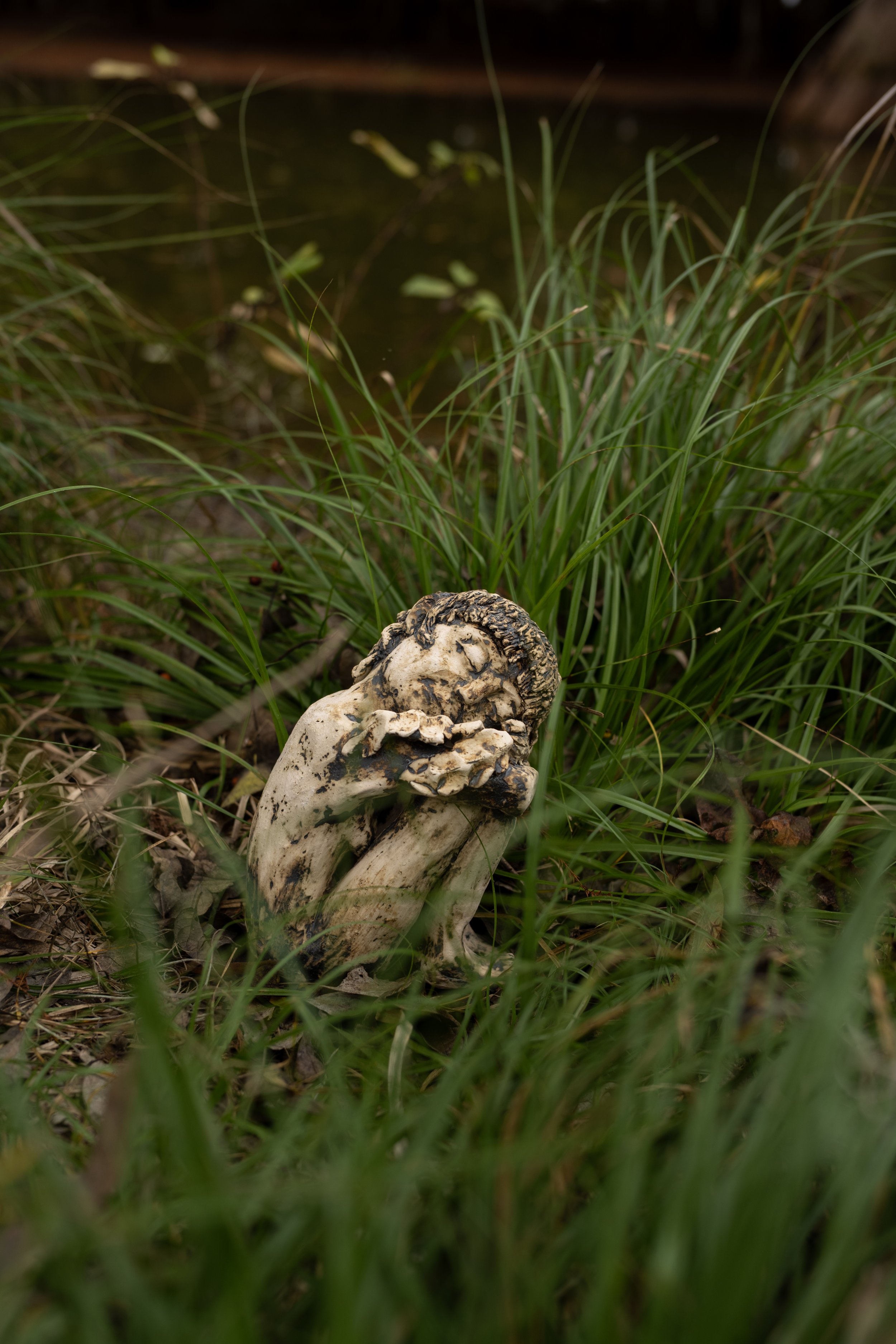 A small statue of a child with curly hair and closed eyes, holding a flower near its face, surrounded by grass.