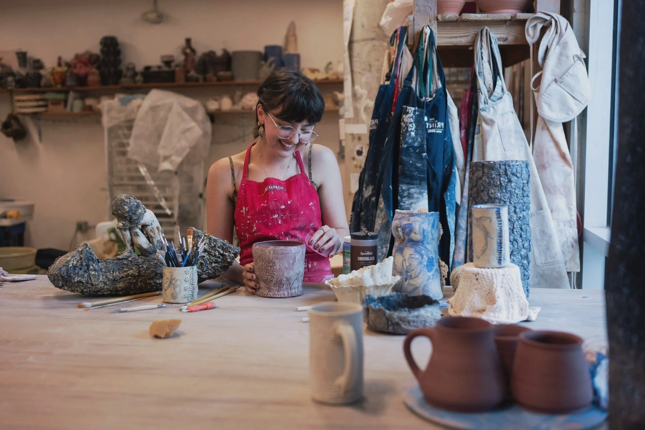 A woman in a red apron is working on ceramic art in a studio, surrounded by various pottery pieces, tools, and art supplies, with shelves and aprons hanging in the background.