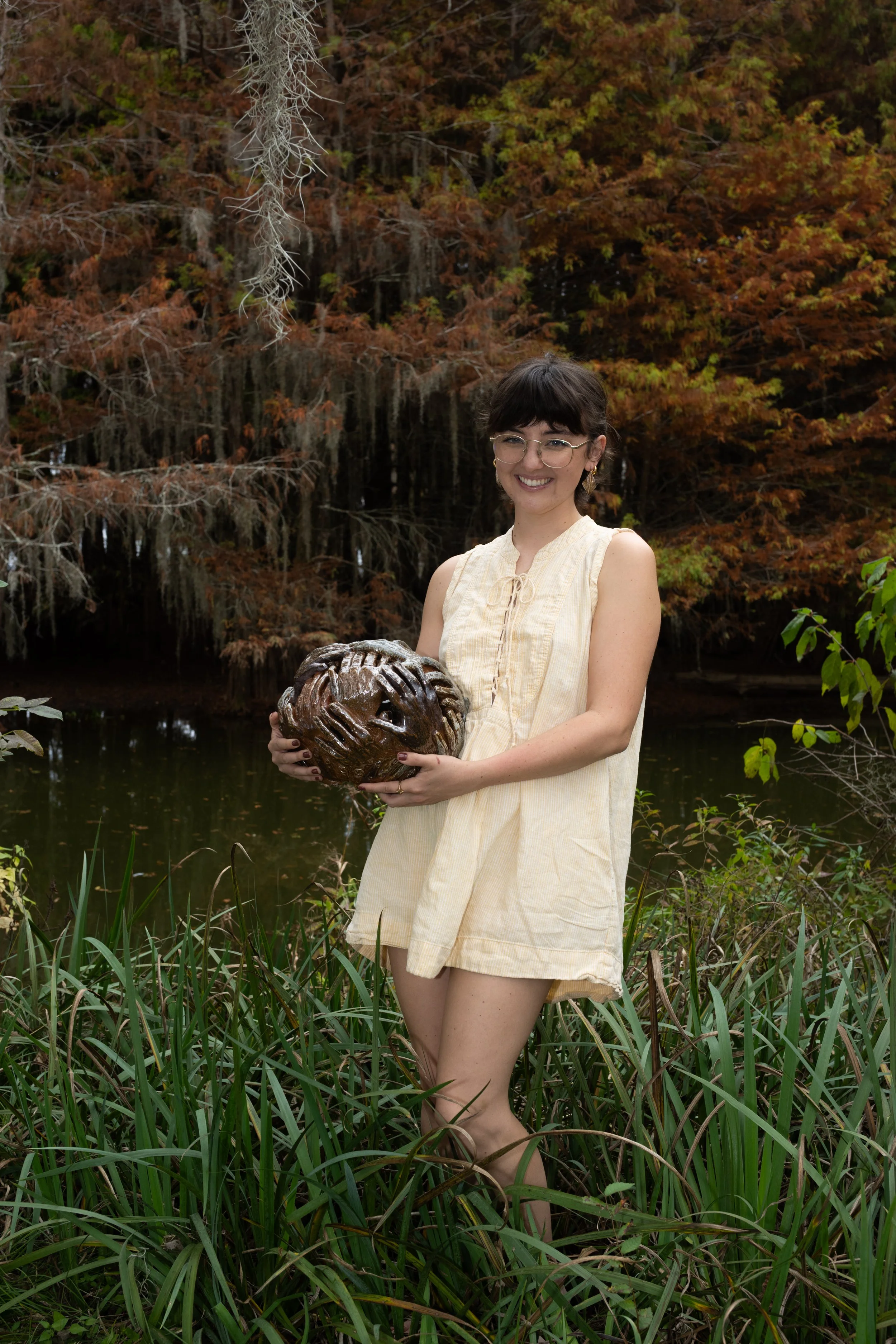 A young woman with dark hair, glasses, and a light yellow sleeveless dress, is standing outdoors in a natural setting with green grass and trees with autumn leaves in the background. She is smiling and holding a carved wooden sphere with hands overlapping, near a body of water.
