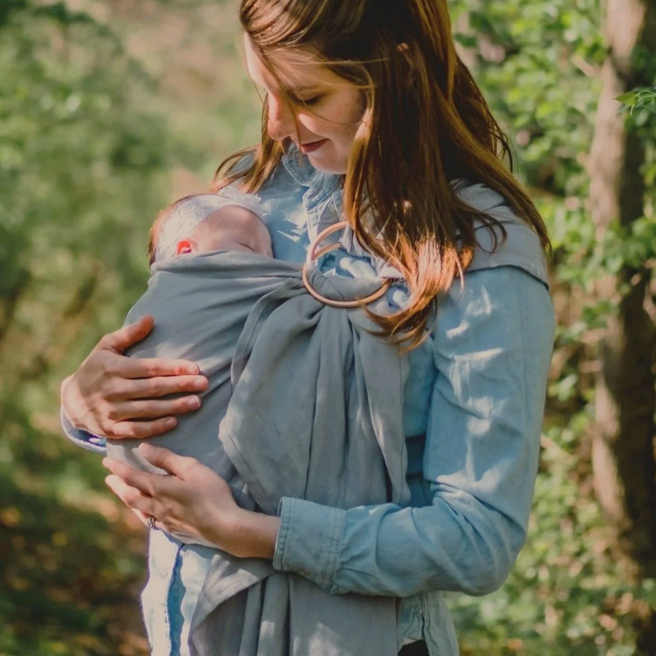 Une femme tient son bébé dans un sling dans ses bras, en extérieur avec des arbres et des feuilles vertes.