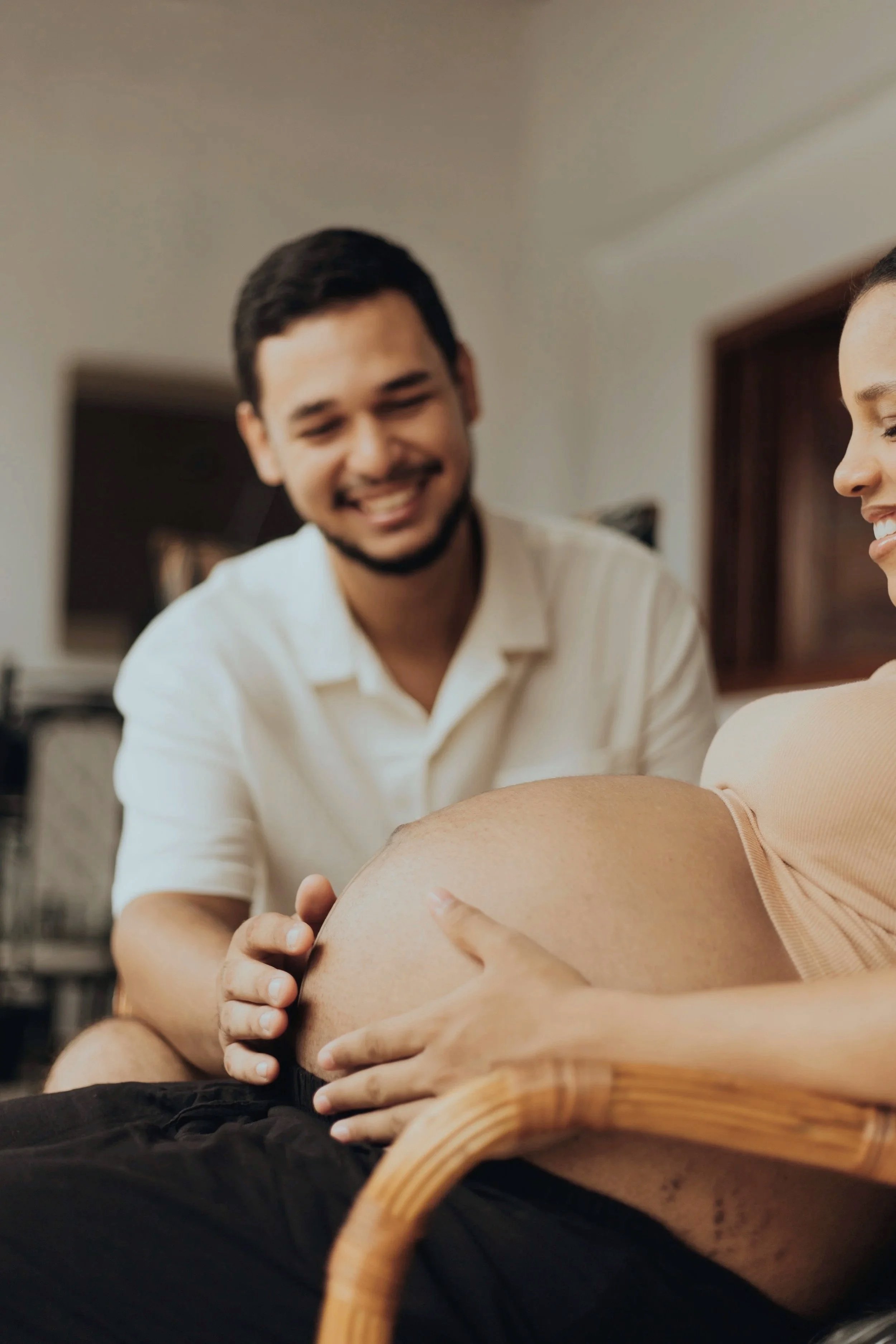 Un homme souriant assis devant une femme enceinte qui touche son ventre. La scène semble se dérouler à l'intérieur, dans une ambiance chaleureuse.