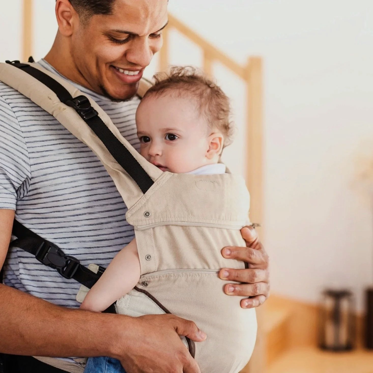 Un homme souriant tient son bébé dans un sac de portage beige, dans une maison.