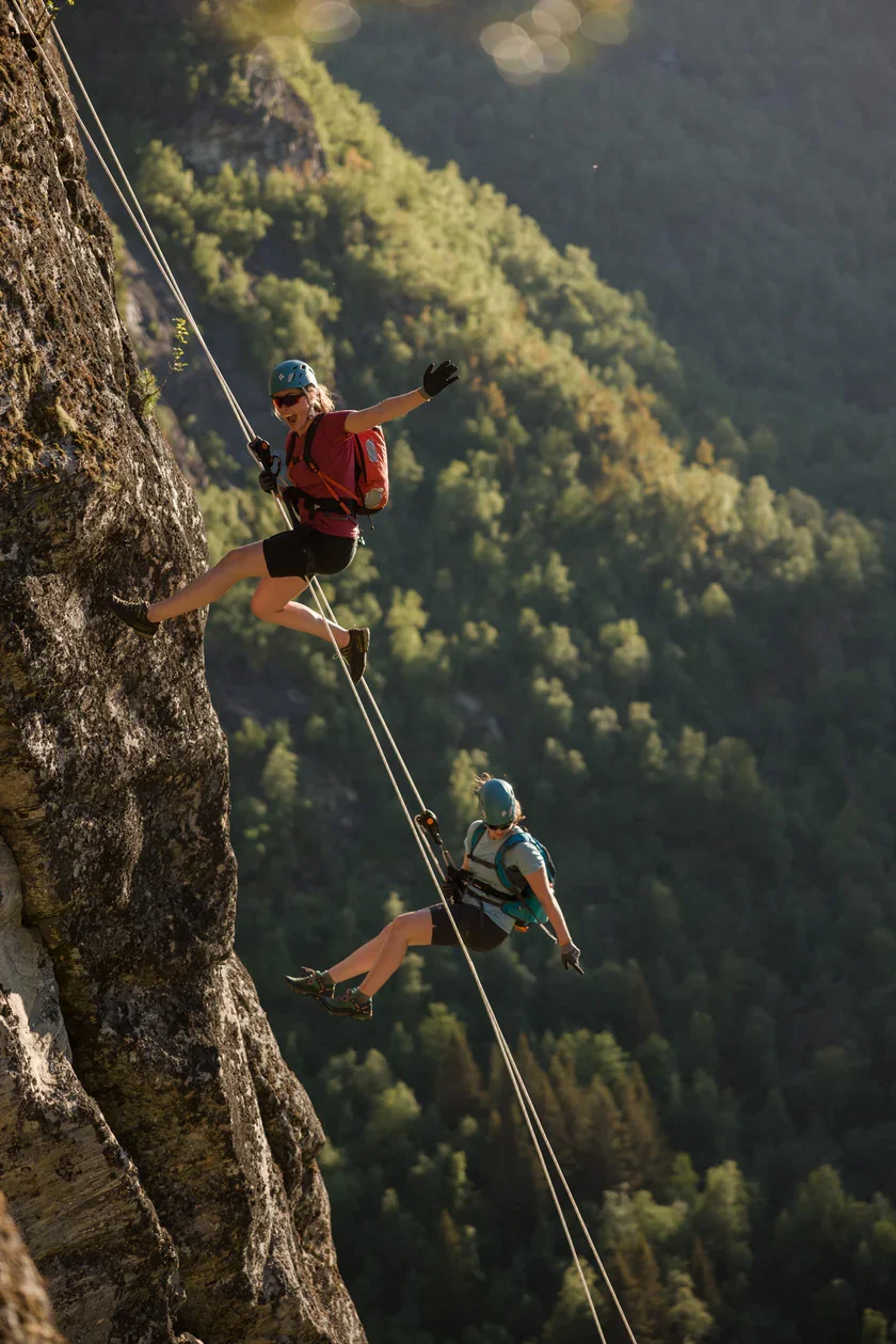 Explore Norway. Two women rock climbing while rappelling down a steep cliff, surrounded by a forested mountain landscape.