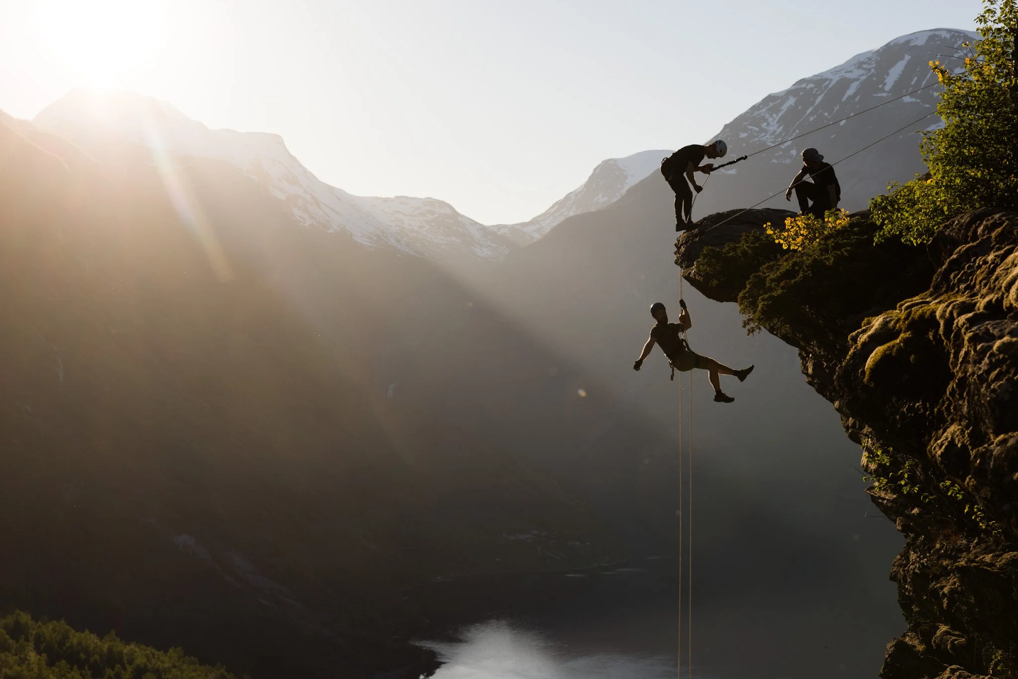 Explore Norway. People rock climbing and rappelling on a cliffside with a mountain and lake in the background during sunset.