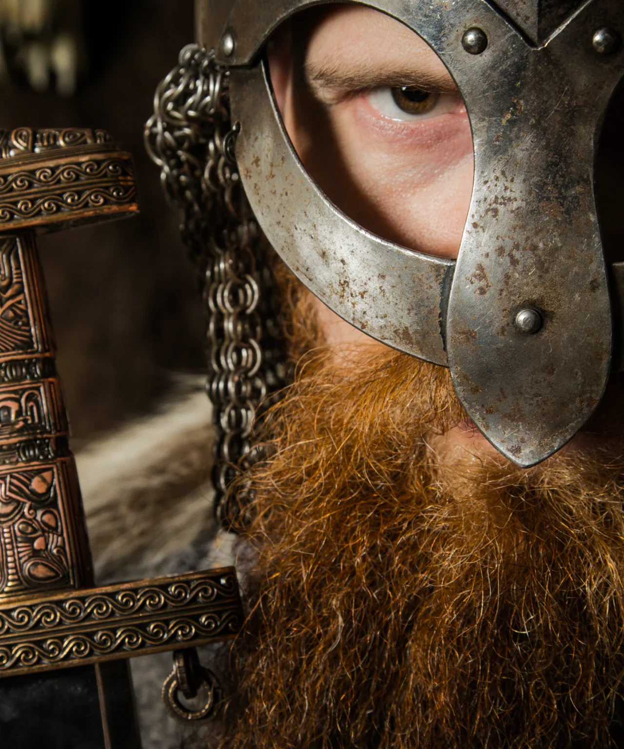 A person wearing a metal helmet with a face opening, a chainmail neck covering, and medieval-style armor, holding a decorative sword.